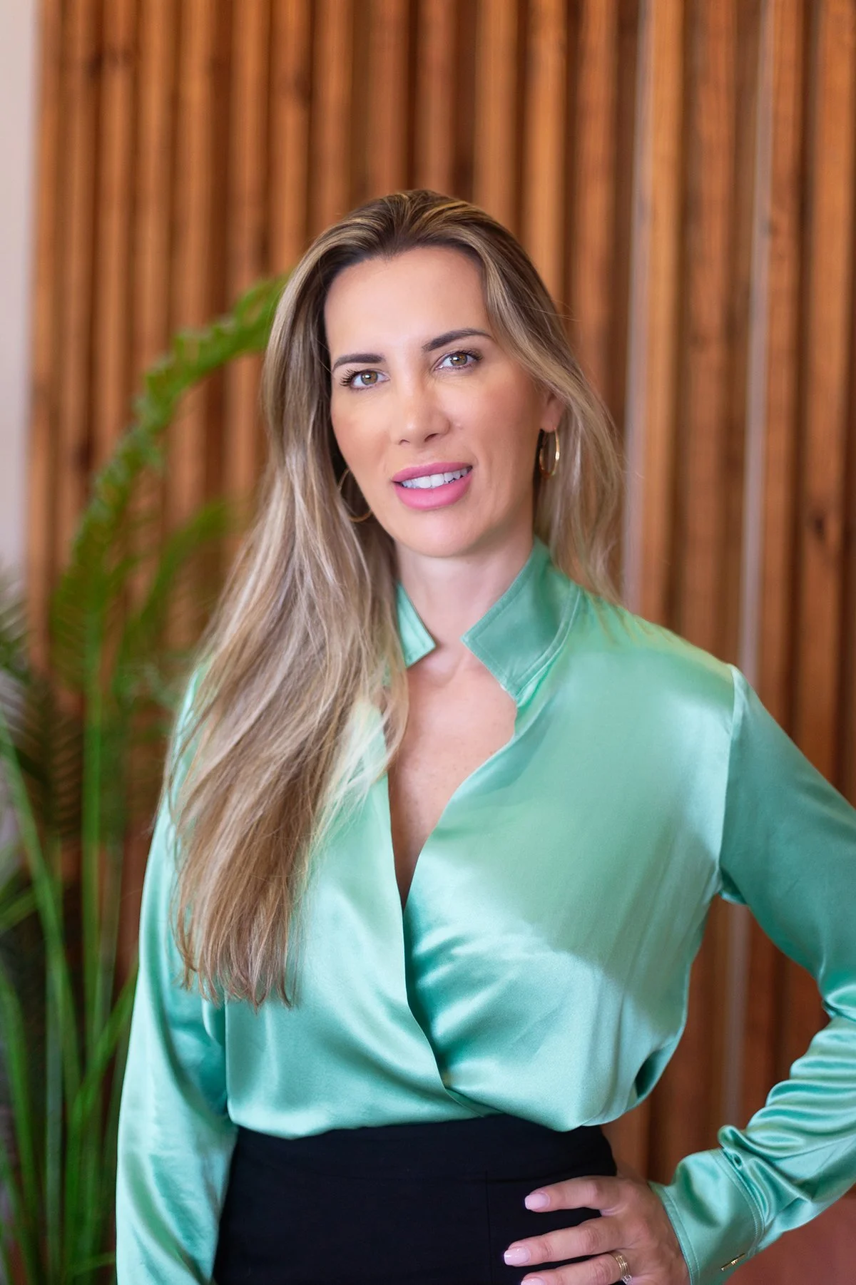 A woman with long, wavy blonde hair wearing a shiny mint green blouse and black pants, standing in front of a wooden background with a green plant on the left.