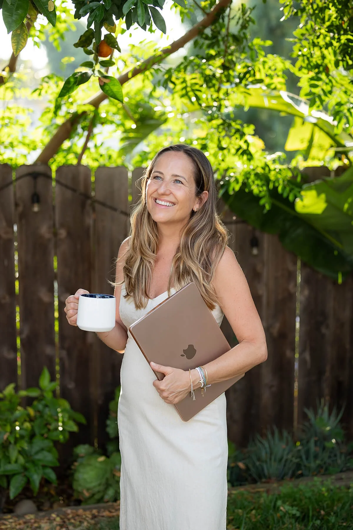 A woman with long hair in a sleeveless white dress holding a white mug and a closed MacBook in a garden with green foliage and a wooden fence, smiling and looking up.