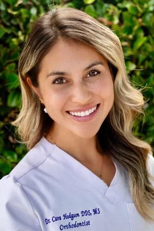Portrait of a smiling woman with wavy hair in front of green foliage, wearing a white dental professional uniform.