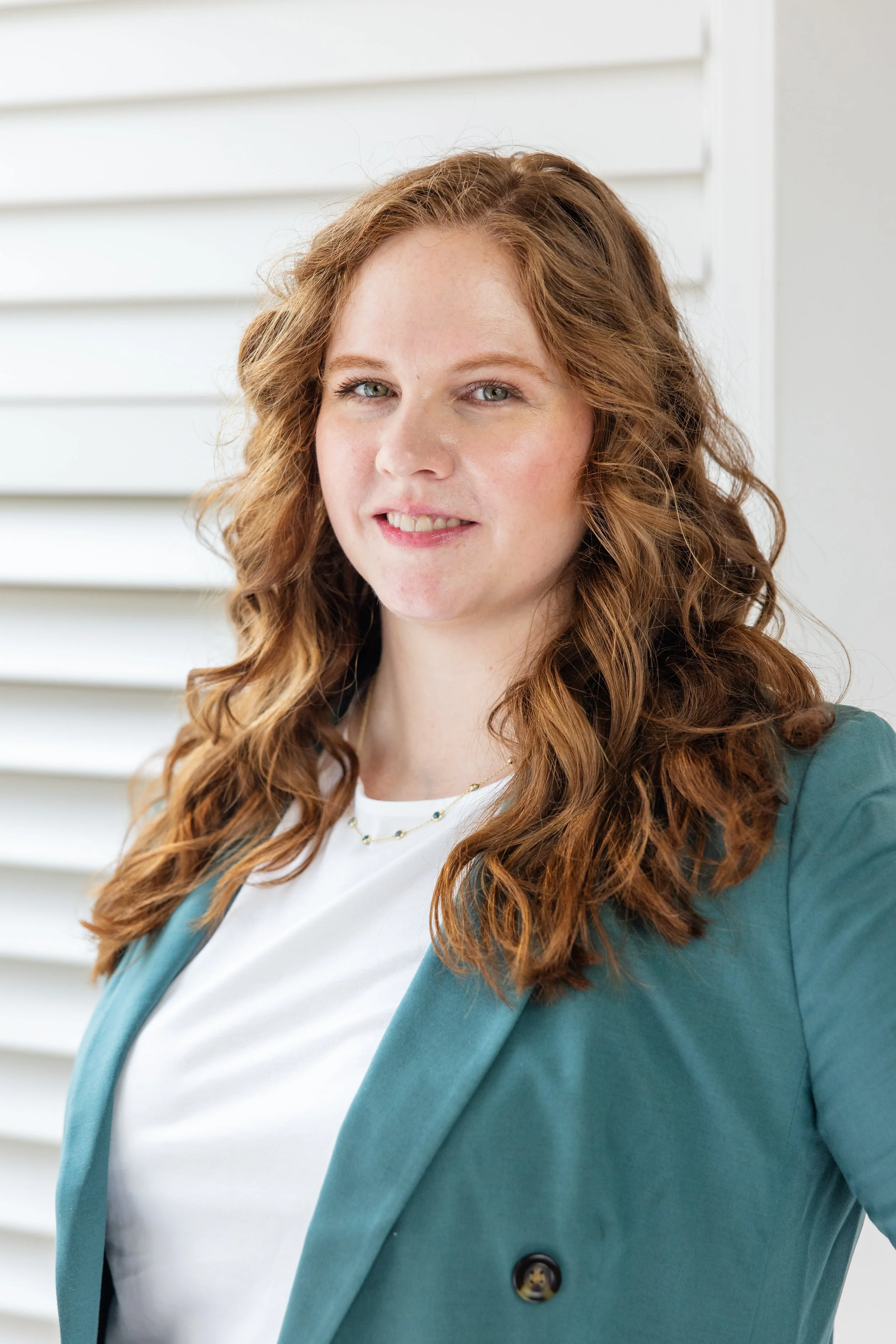 A woman with red curly hair wearing a white top and teal blazer, standing in front of white background with horizontal panels.