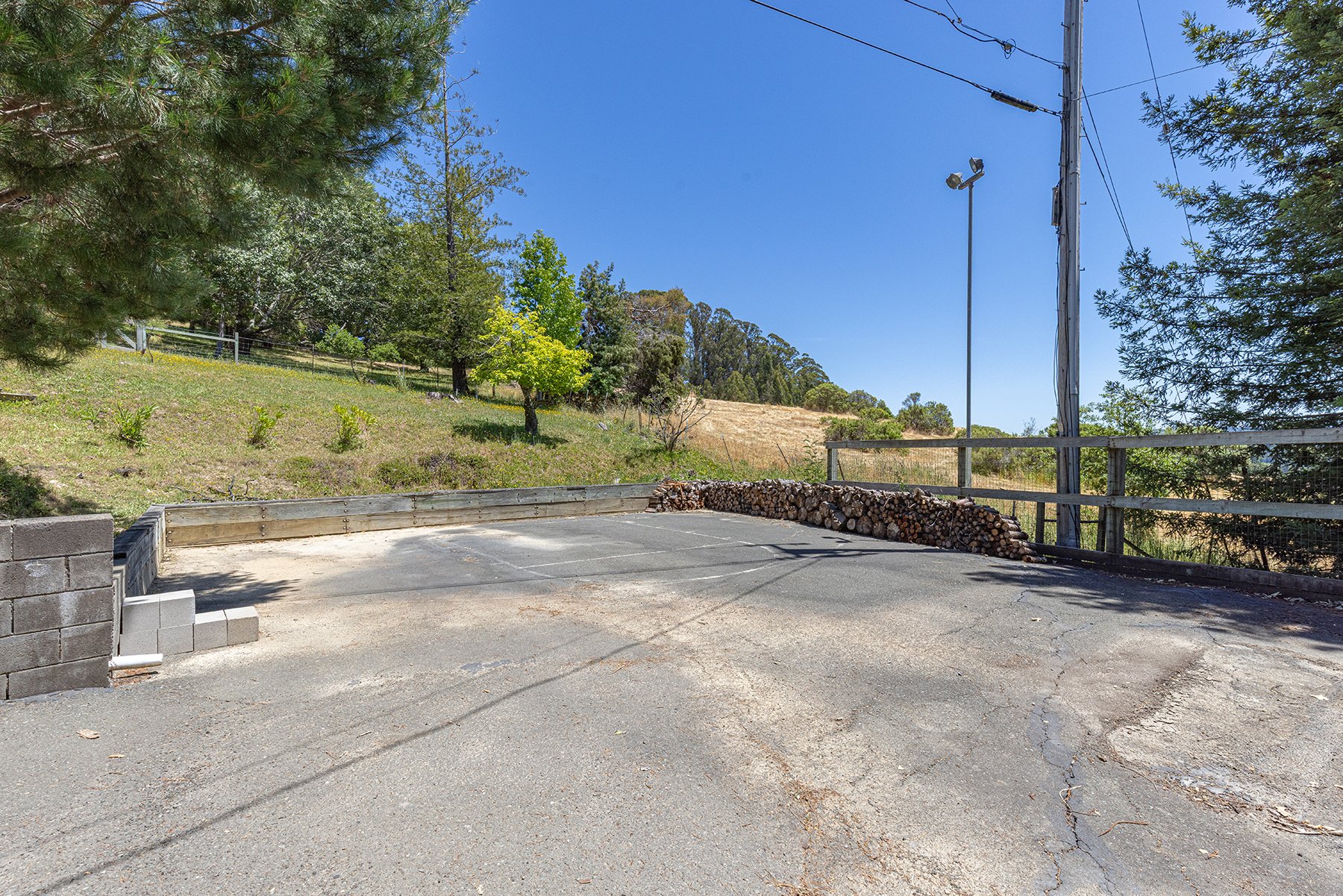 Empty parking lot with a small stack of firewood, wooden fence, trees, and utility pole under a clear blue sky.