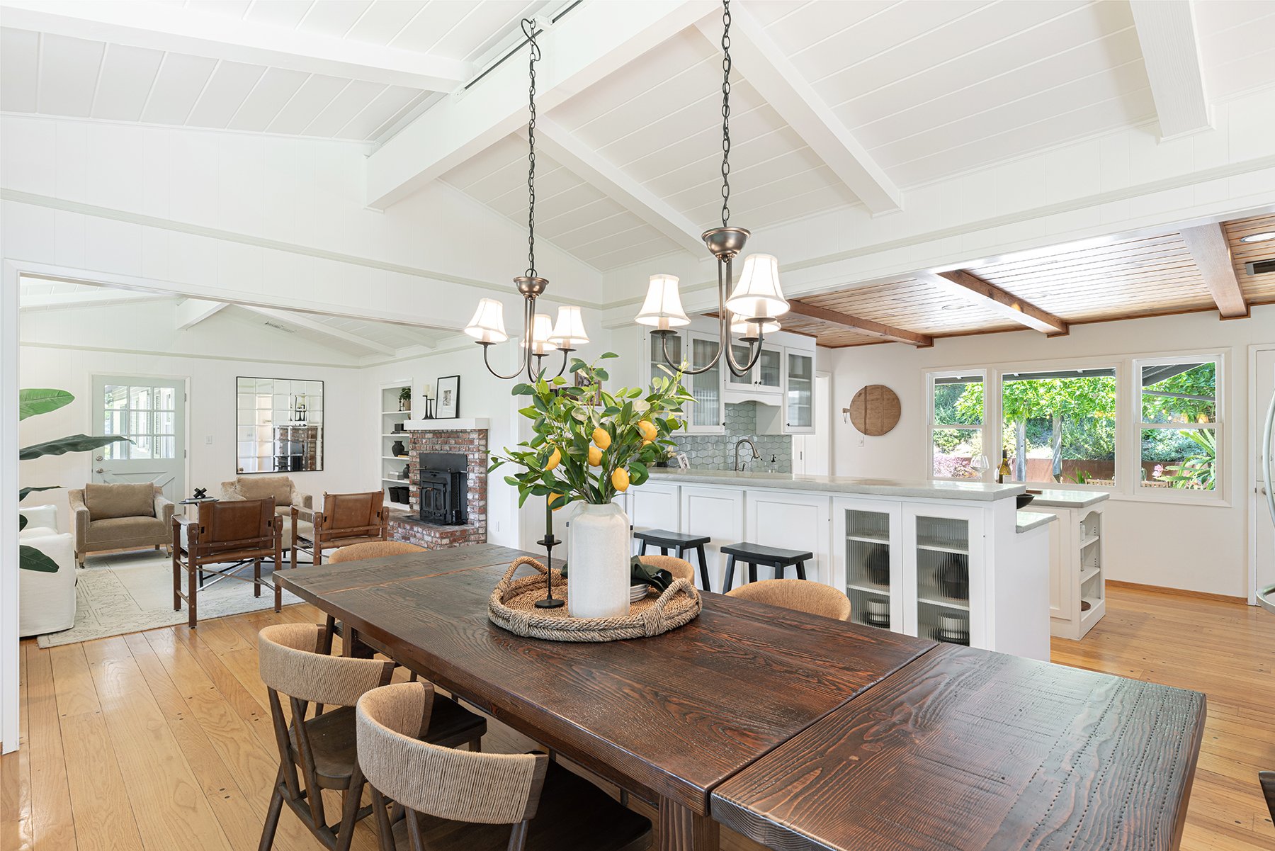 Open-concept kitchen and living room with white walls and ceiling, wooden flooring, a dark wooden dining table with a white vase and lemon branch centerpiece, and a cozy living area with beige and brown seating, a brick fireplace, and large windows l