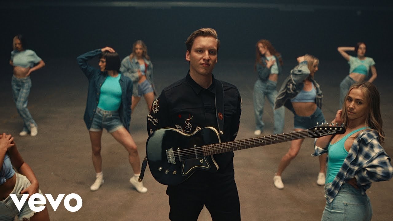 George Ezra holding a guitar in a dance studio with nine women, including, Crystal Leah Jones in casual clothing standing or posing in the background.