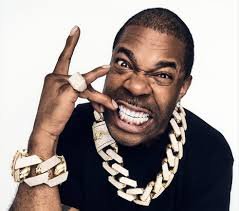 A man making a playful face, showing his teeth, wearing multiple gold chains and a large ring, with his hand near his face in a gesture.