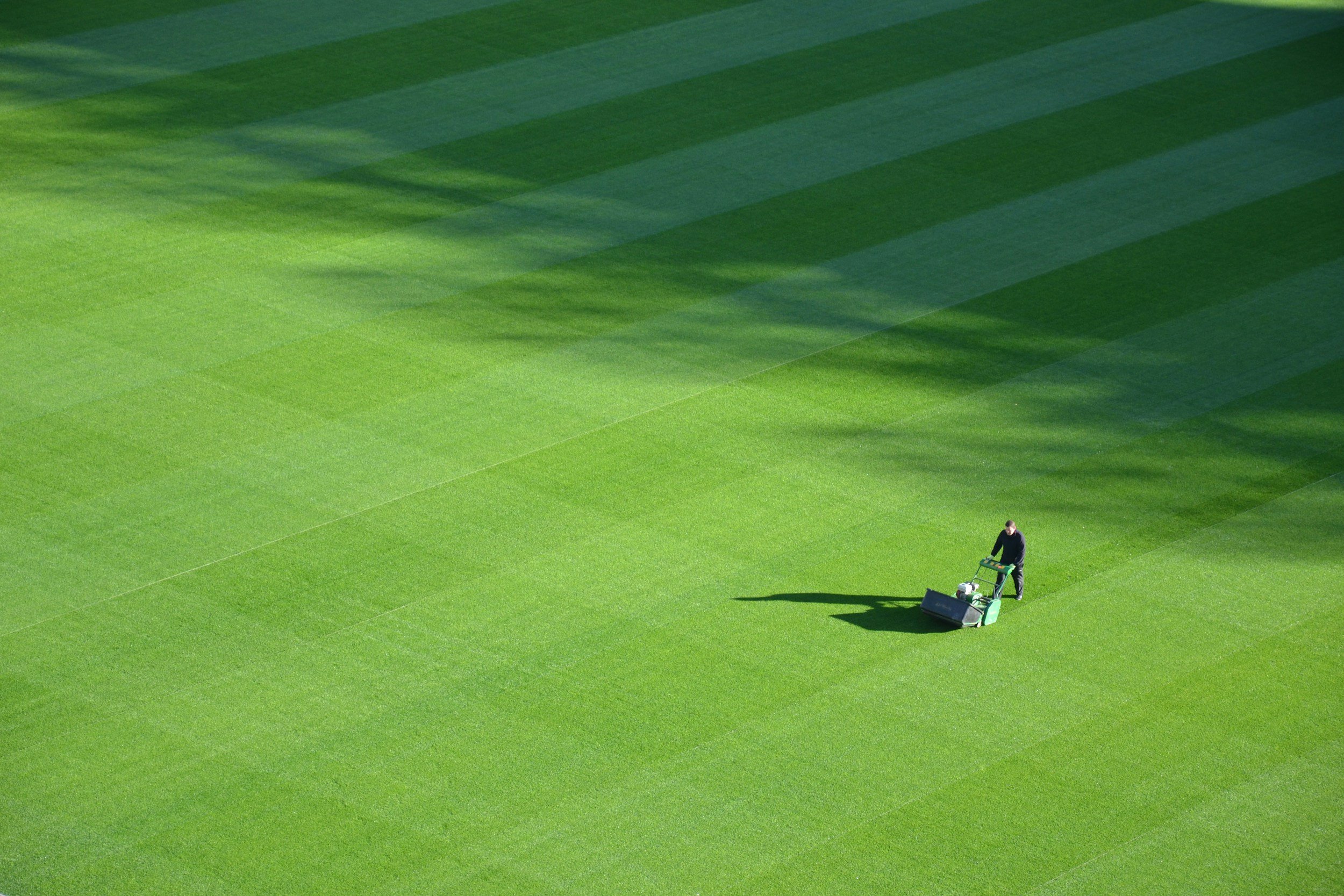 A person is using a lawn mower on a large, well-maintained green grass field, with shadows cast by trees or structures overhead.