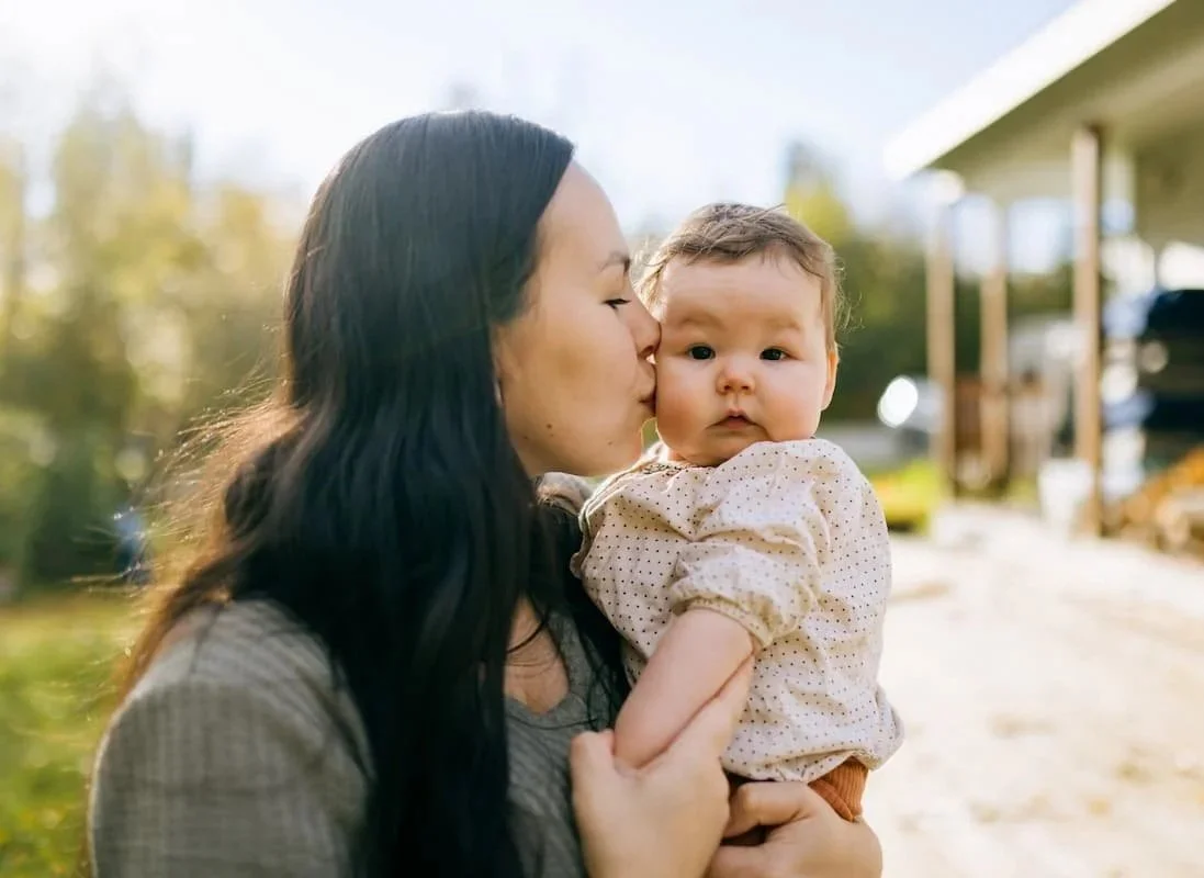 A woman with long black hair holding and kissing a baby girl on the cheek outdoors on a sunny day.