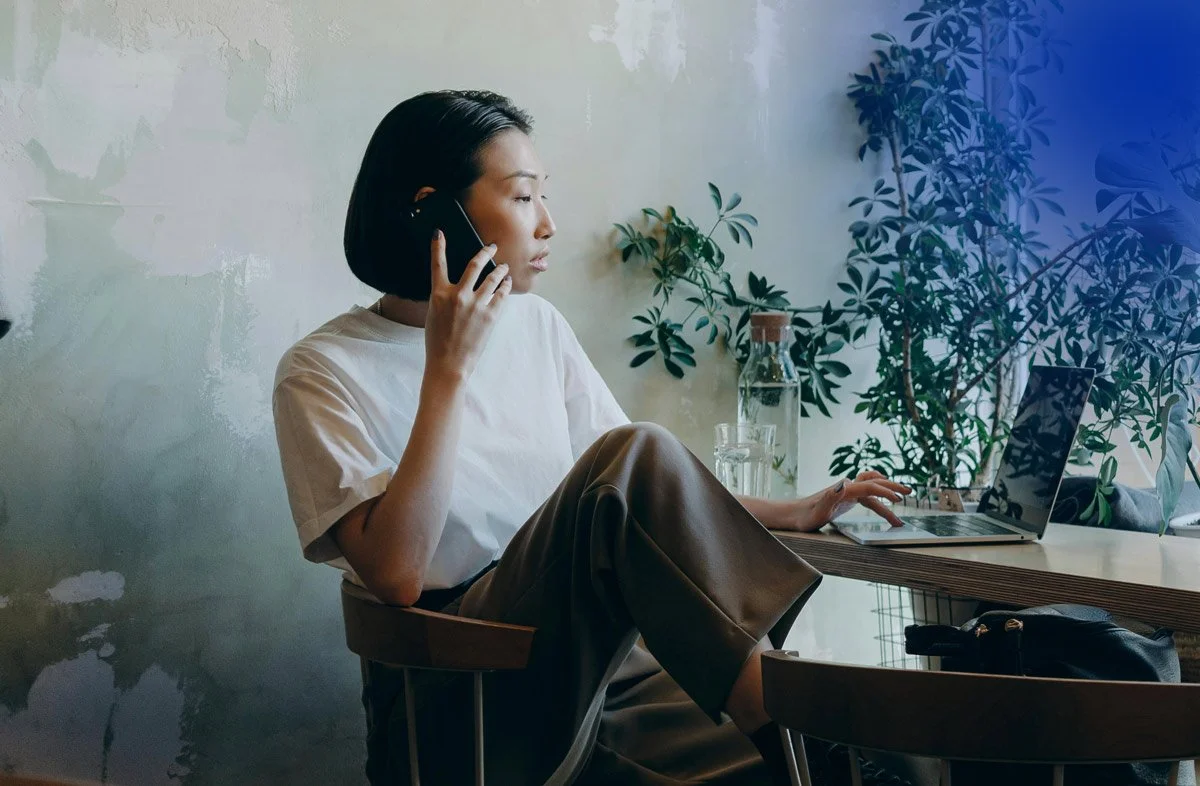 Image of woman sitting in a cafe taking a call in front of her computer.
