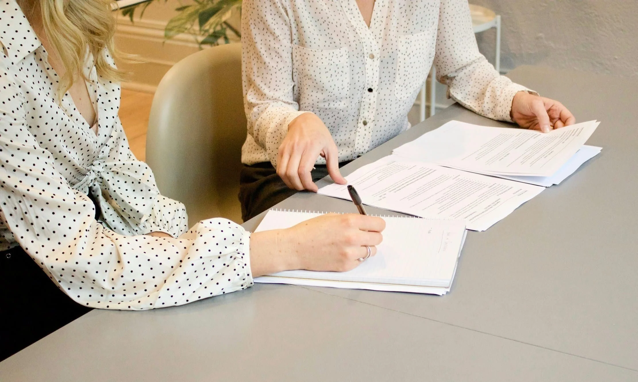 Two women sitting at a table with papers, one writing in a notebook and the other pointing at a document.