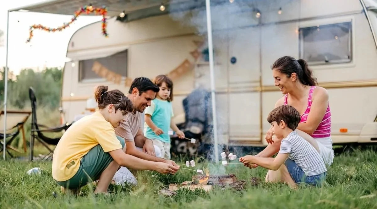 Family roasting marshmallows over a campfire outside a camper trailer with string lights and autumn decorations.
