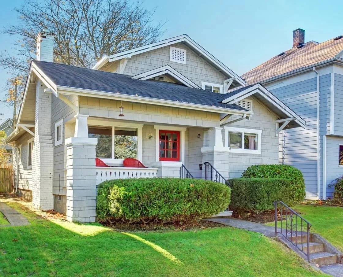 A white, two-story house with a front porch, red door, and well-maintained green lawn and shrubs