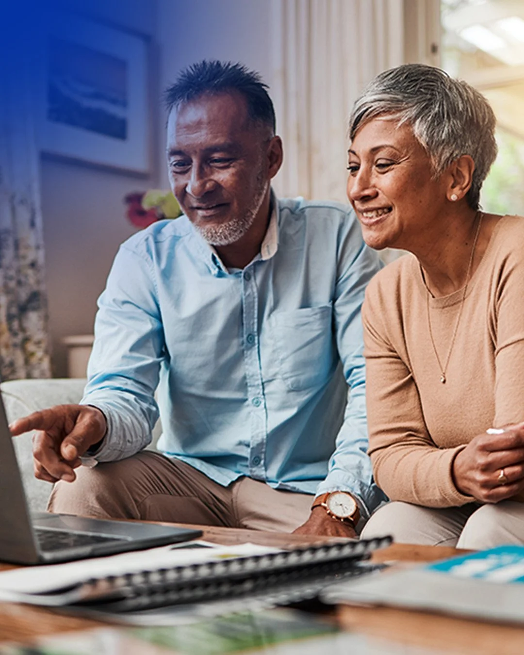 An elderly man and woman sitting together, looking at a laptop and smiling in a well-lit room.
