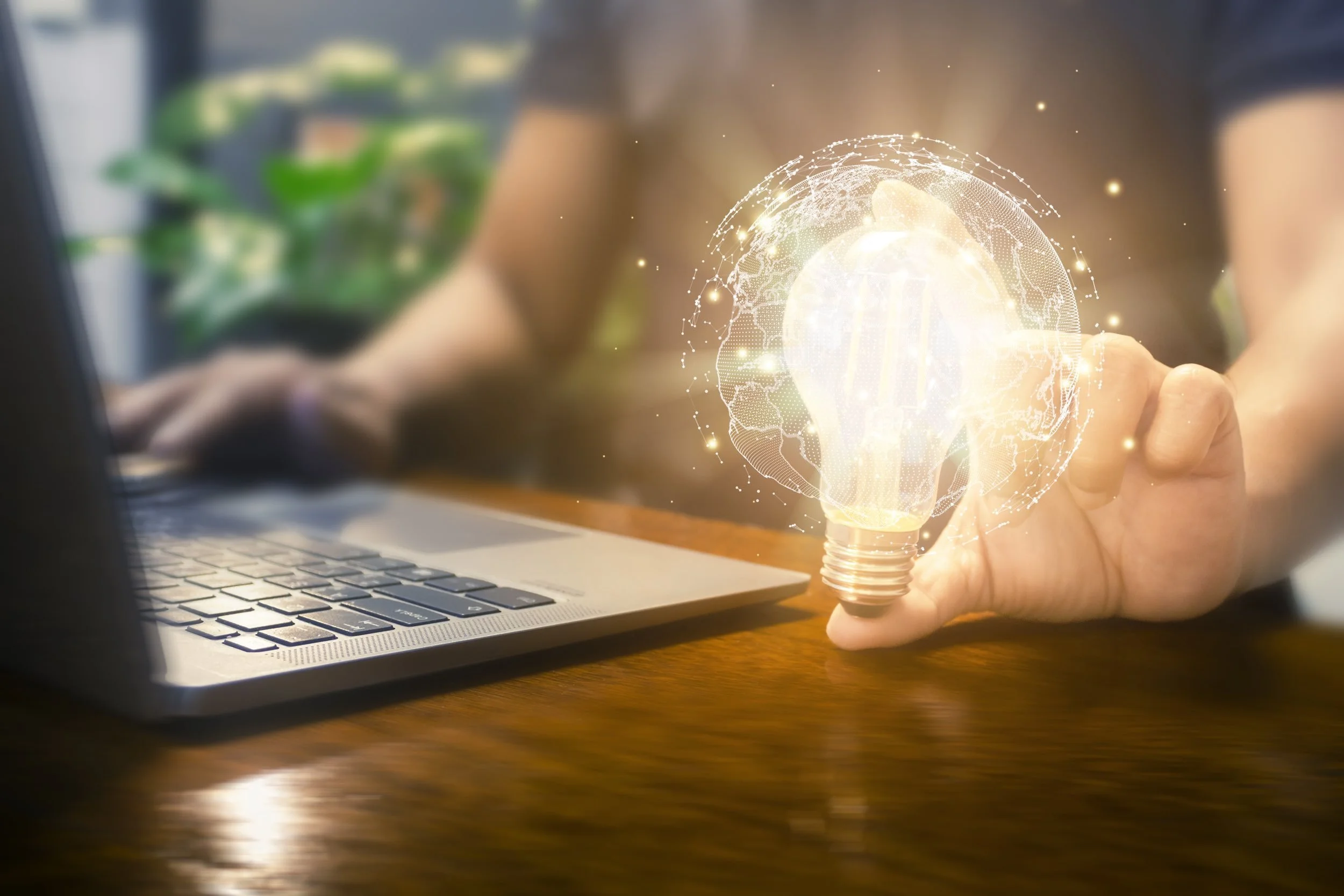 Person using a laptop with a digital lightbulb and world globe hologram in the foreground.