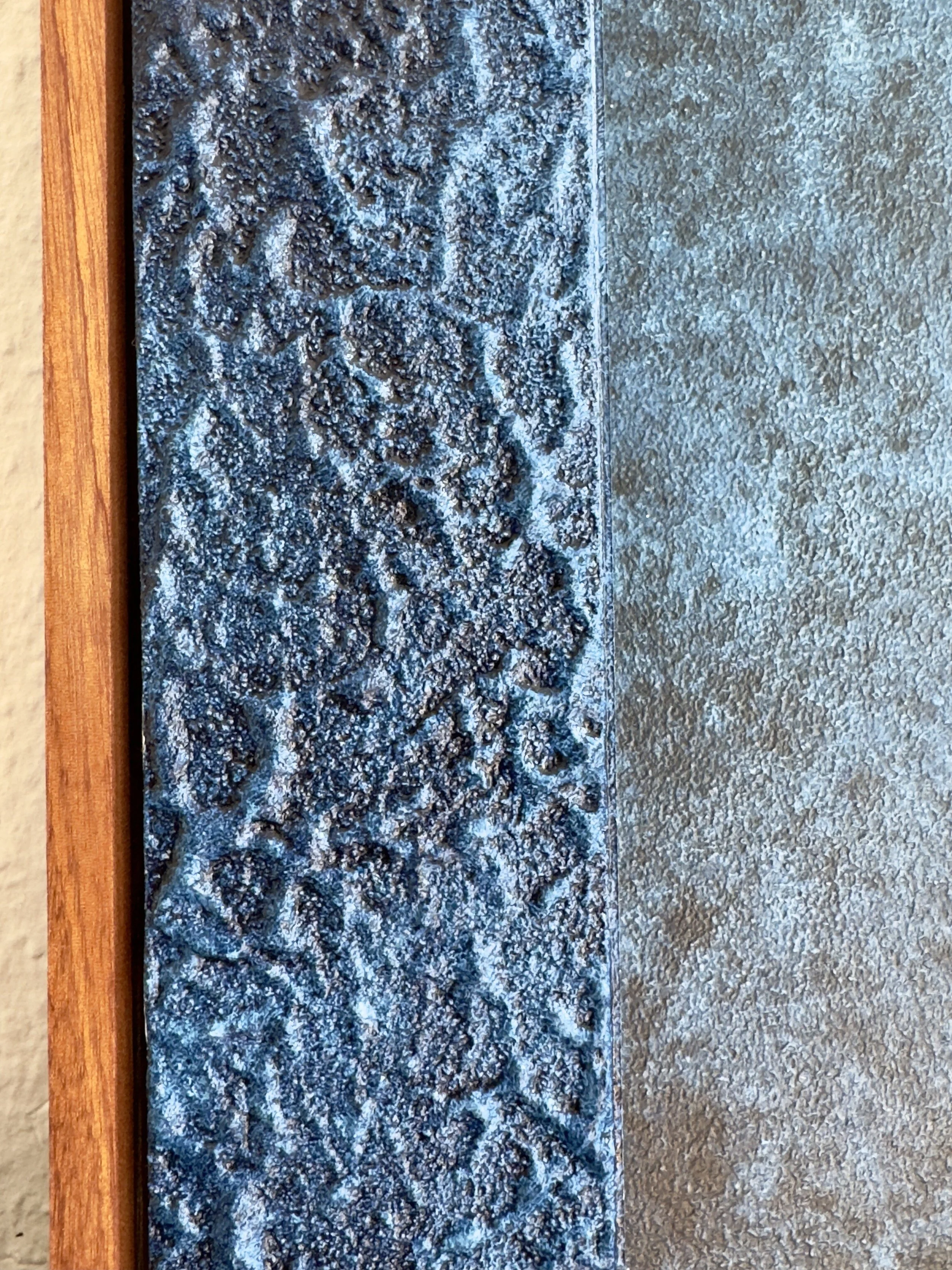 Ein dekoratives schmiedeeisernes Element mit floralem Muster, eingerahmt von Holz auf der linken Seite und einer hellgrauen Wand auf der rechten Seite.