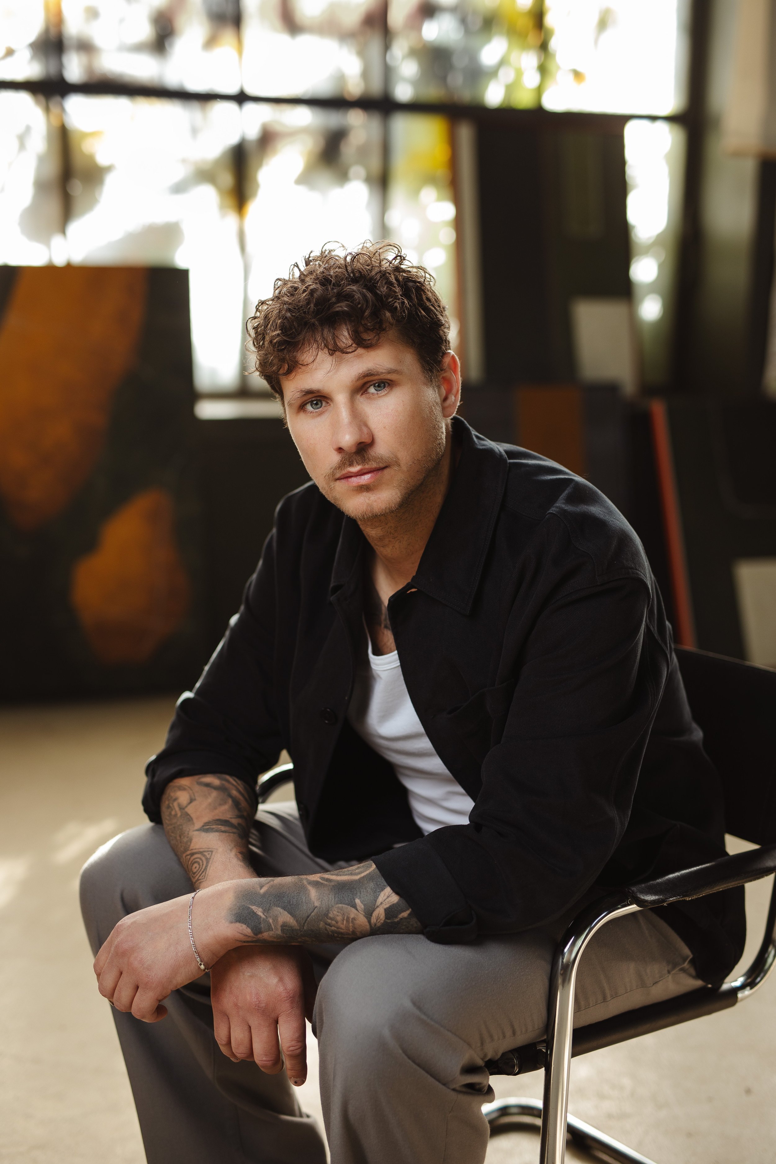 Junger Mann mit Tattoos sitzt in einem modernen Raum, blickt nach vorne und trägt eine schwarze Jacke, weiße T-Shirt und graue Hosen.