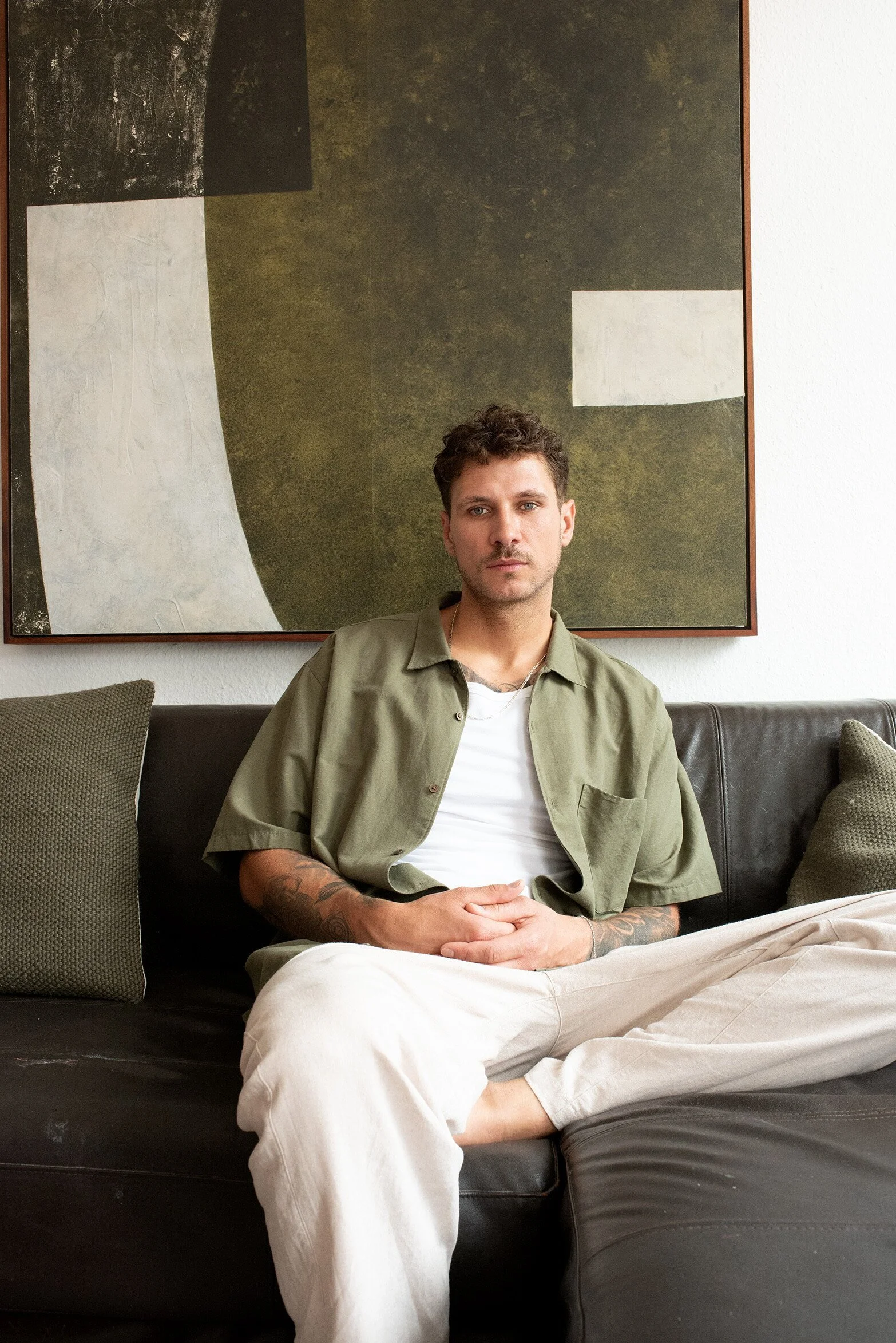 Ein junger Mann mit Tattoos sitzt entspannt auf einem schwarzen Sofa vor einer abstrakten Wandkunst. Er trägt eine offene grüne Jacke über einem weißen T-Shirt und beige Hosen.