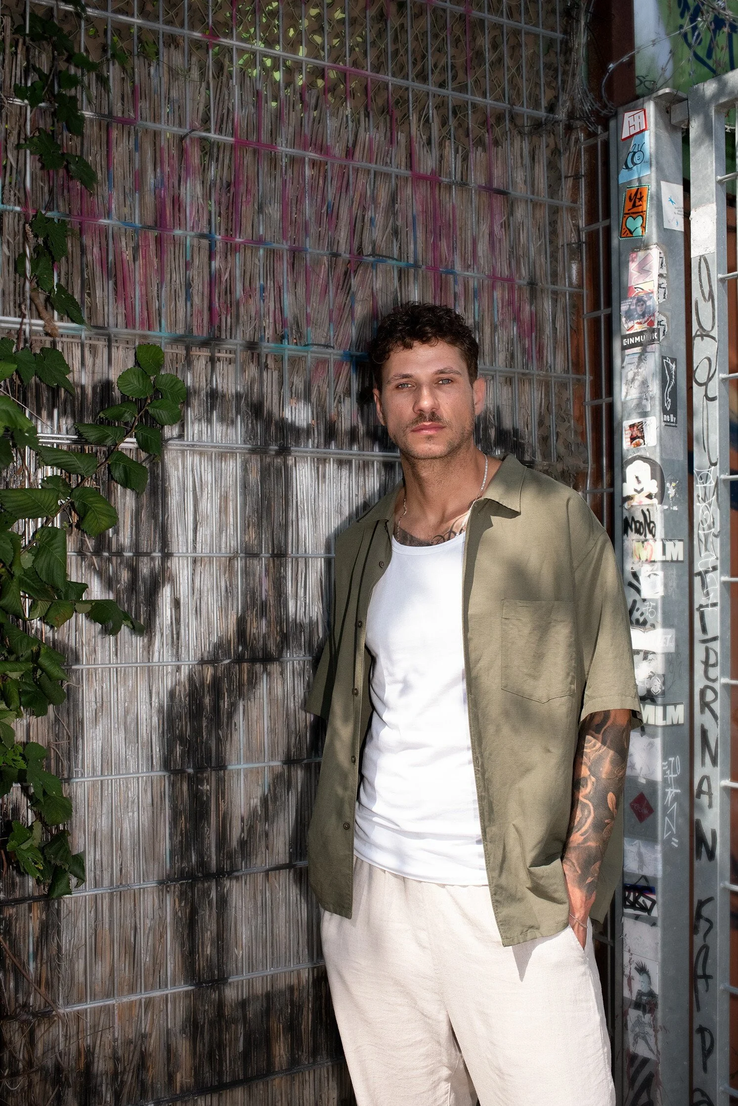 Ein junger Mann mit dunklen, lockigen Haaren steht vor einer Metallabsperrung mit Graffiti, trägt ein grünes Hemd, ein weißes T-Shirt und beige Hosen, und blickt ernst in die Kamera.