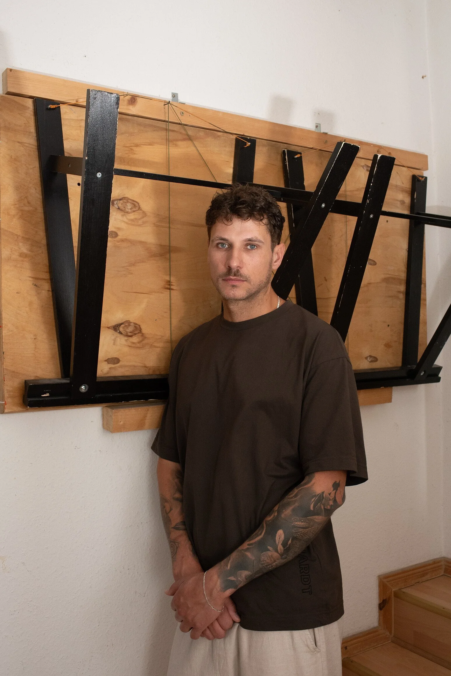 Ein junger Mann mit dunklem T-Shirt und Tattoos auf den Armen steht vor einer Wand mit Holzrahmen und schwarzen Metallstangen.