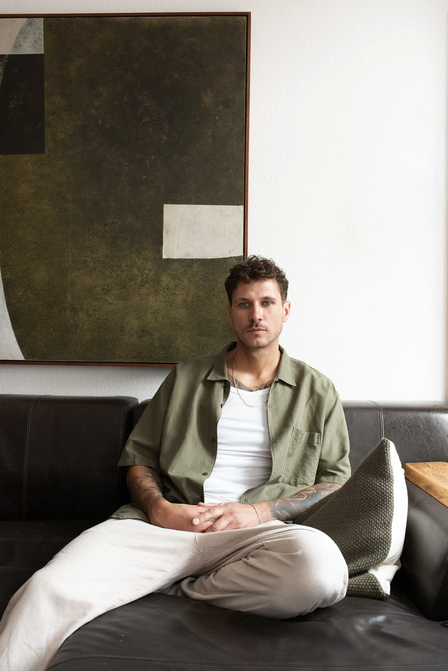 Junger Mann sitzt entspannt auf einem schwarzen Sofa mit weißen Hosen, einem grünen Hemd (offen) und einem weißen T-Shirt, vor einem weißen Wand mit einem großen abstrakten Kunstwerk.