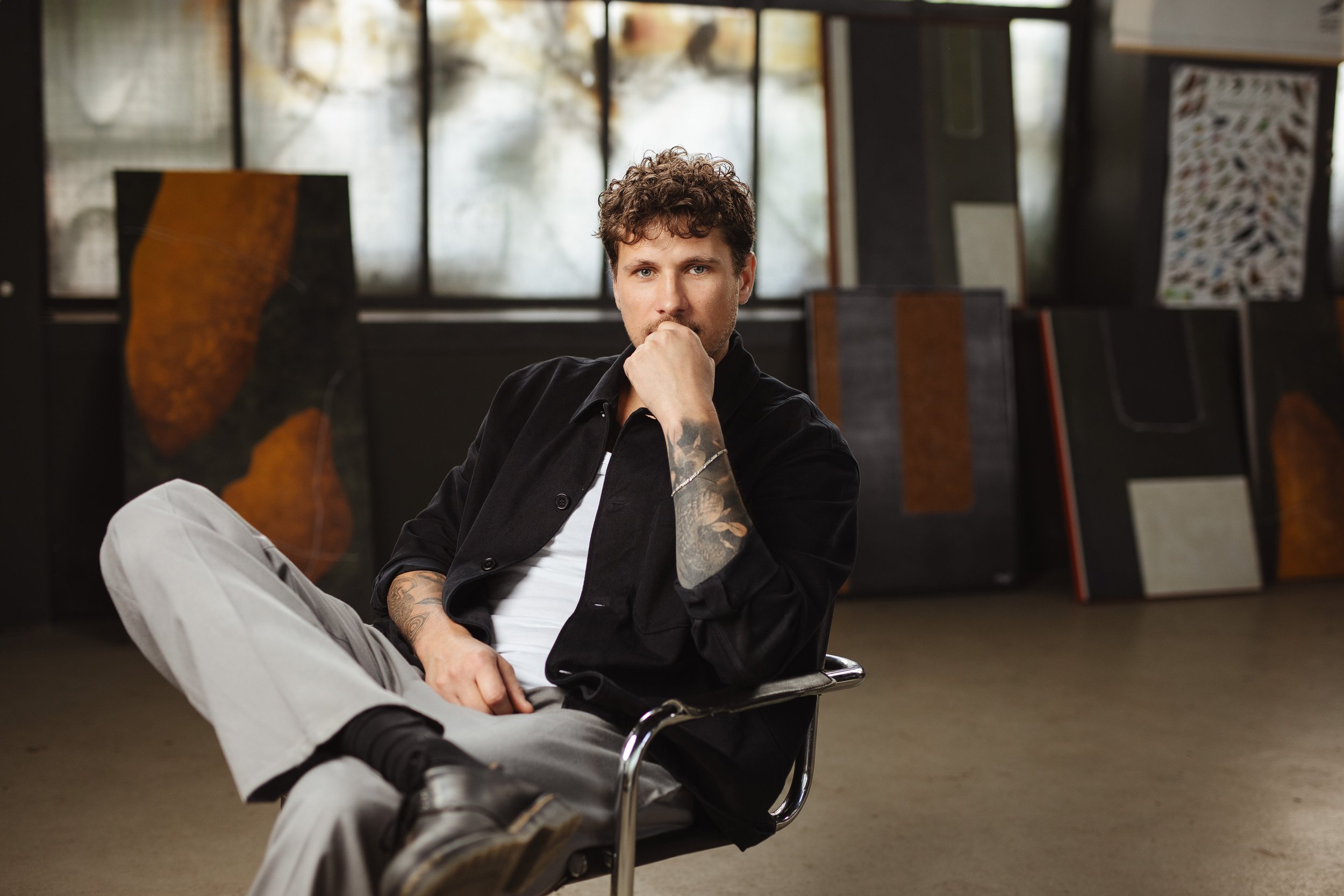 Ein junger Mann mit braunen, lockigen Haaren, Tattoos am Arm, sitzend in einem modernen Raum mit großem Fenster und Kunstwerken im Hintergrund, schaut nach vorne.