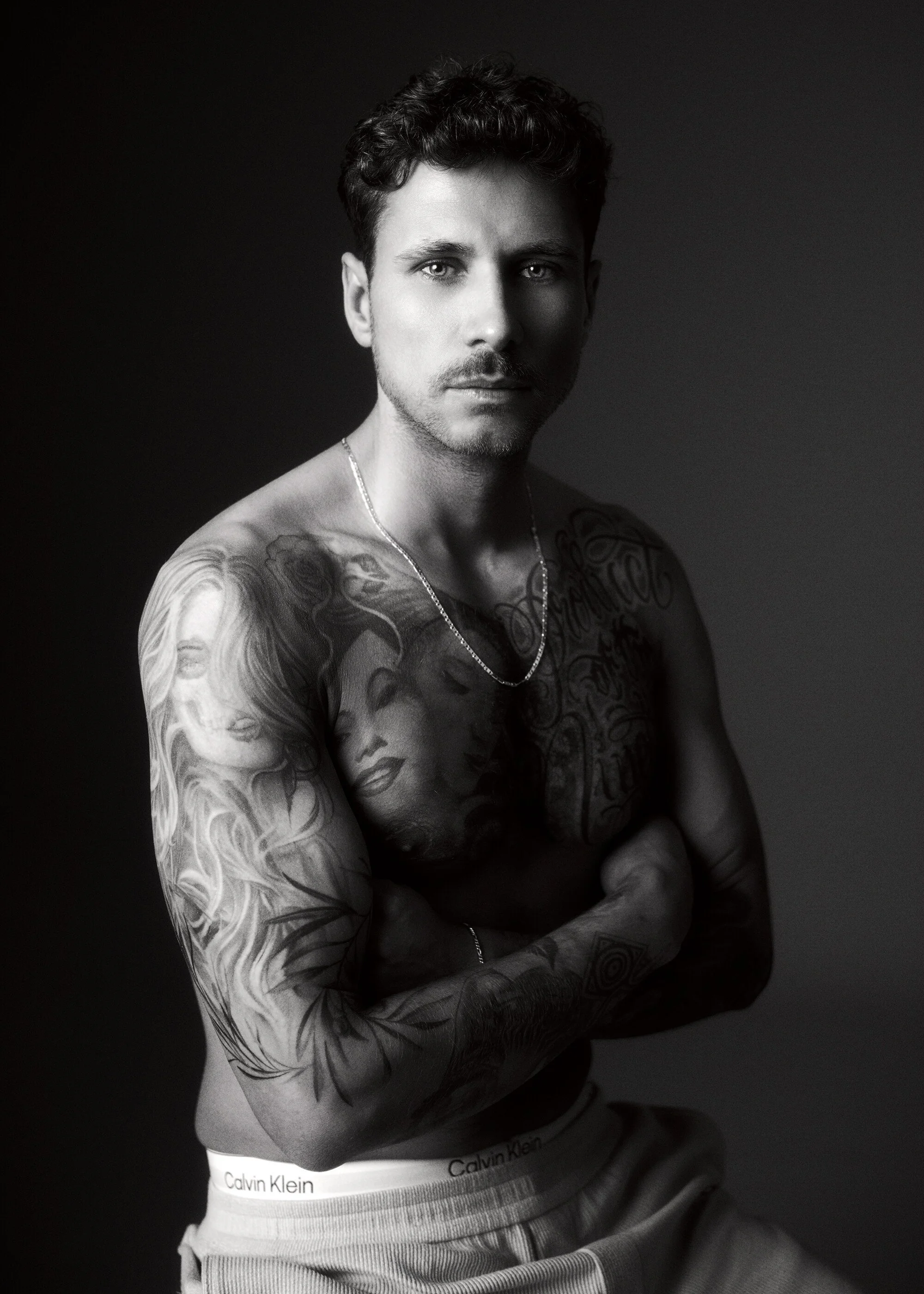 Schwarze und weiße Aufnahme eines jungen Mannes mit Tattoos auf Armen und Brust, kurze dunkle Haare, Blick in die Kamera, no shirt, mit Calvin Klein Unterwäsche, gegen dunklen Hintergrund.