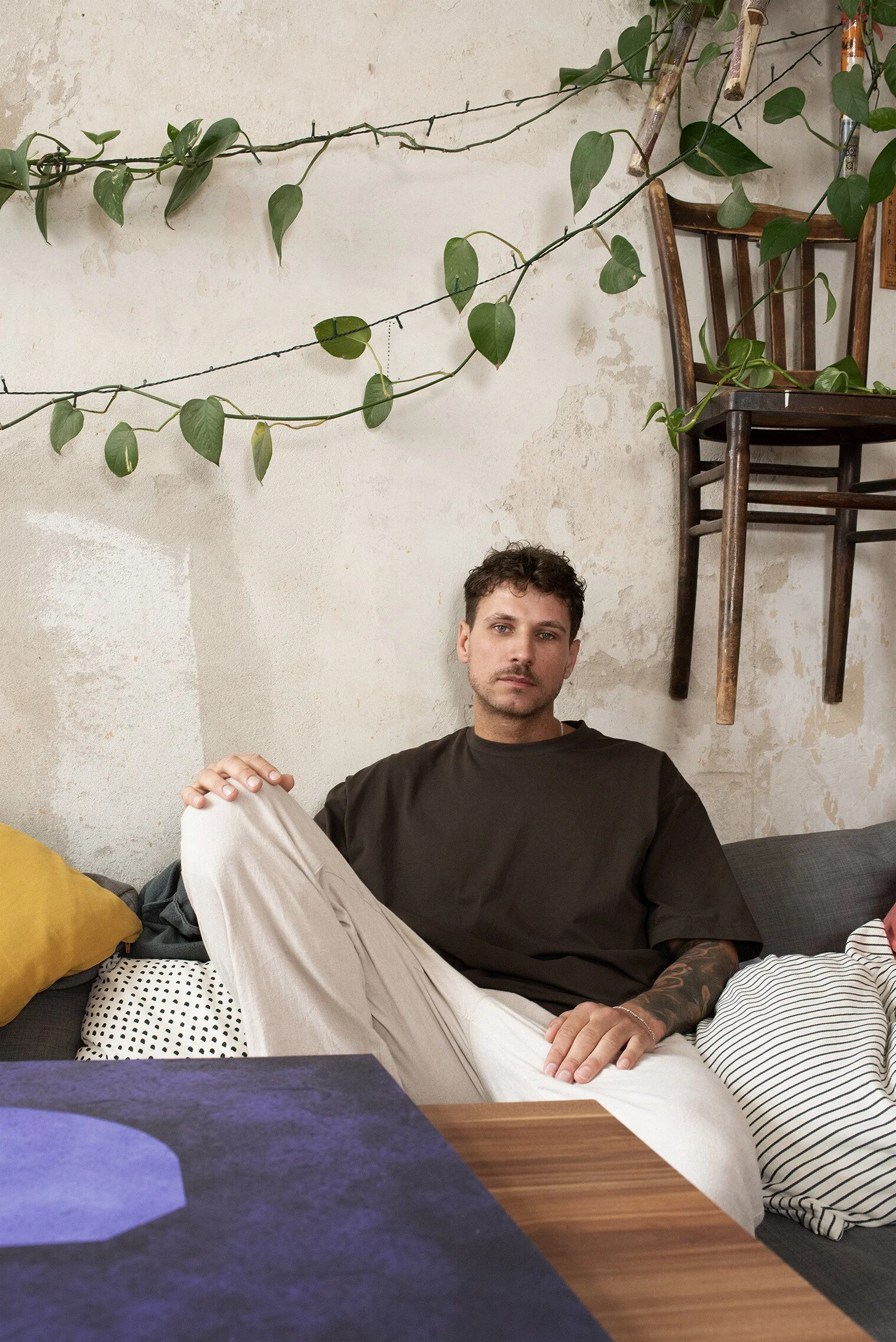 Ein junger Mann sitzt entspannt auf einer Couch in einem Raum mit einer weißen Wand und großen grünen Pflanzen. Er trägt ein schwarzes T-Shirt und beige Hosen.