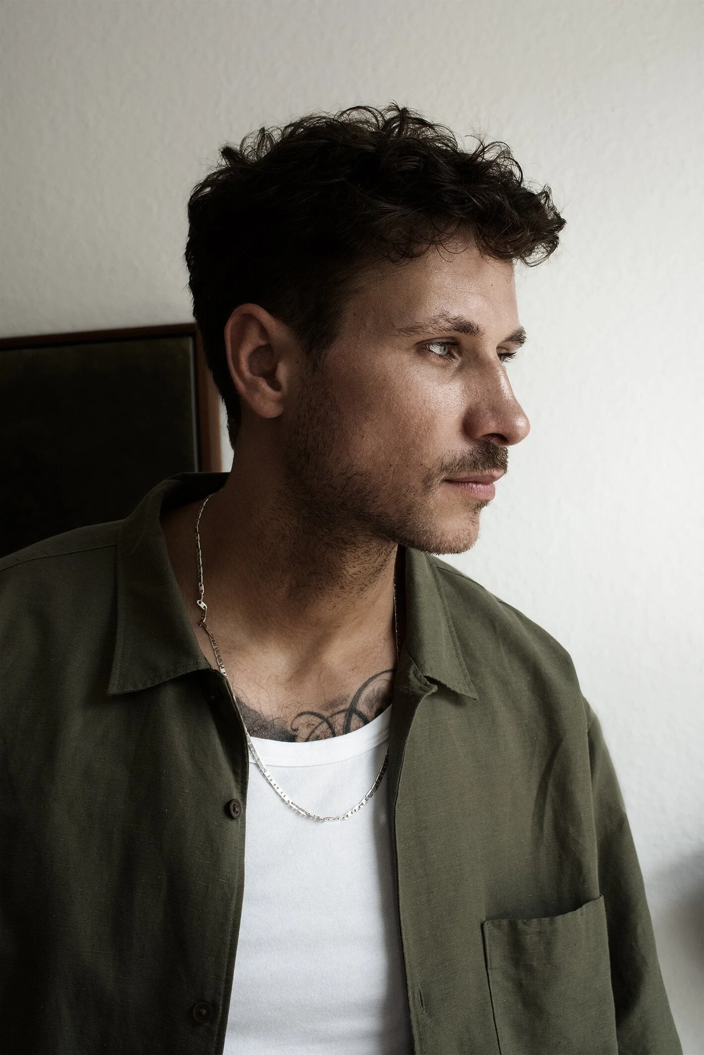 Porträt eines jungen Mannes mit dunklem, lockigem Haar, Bart und Tattoos, trägt einen weißen T-Shirt und eine grüne Jacke, vor neutralem Hintergrund.
