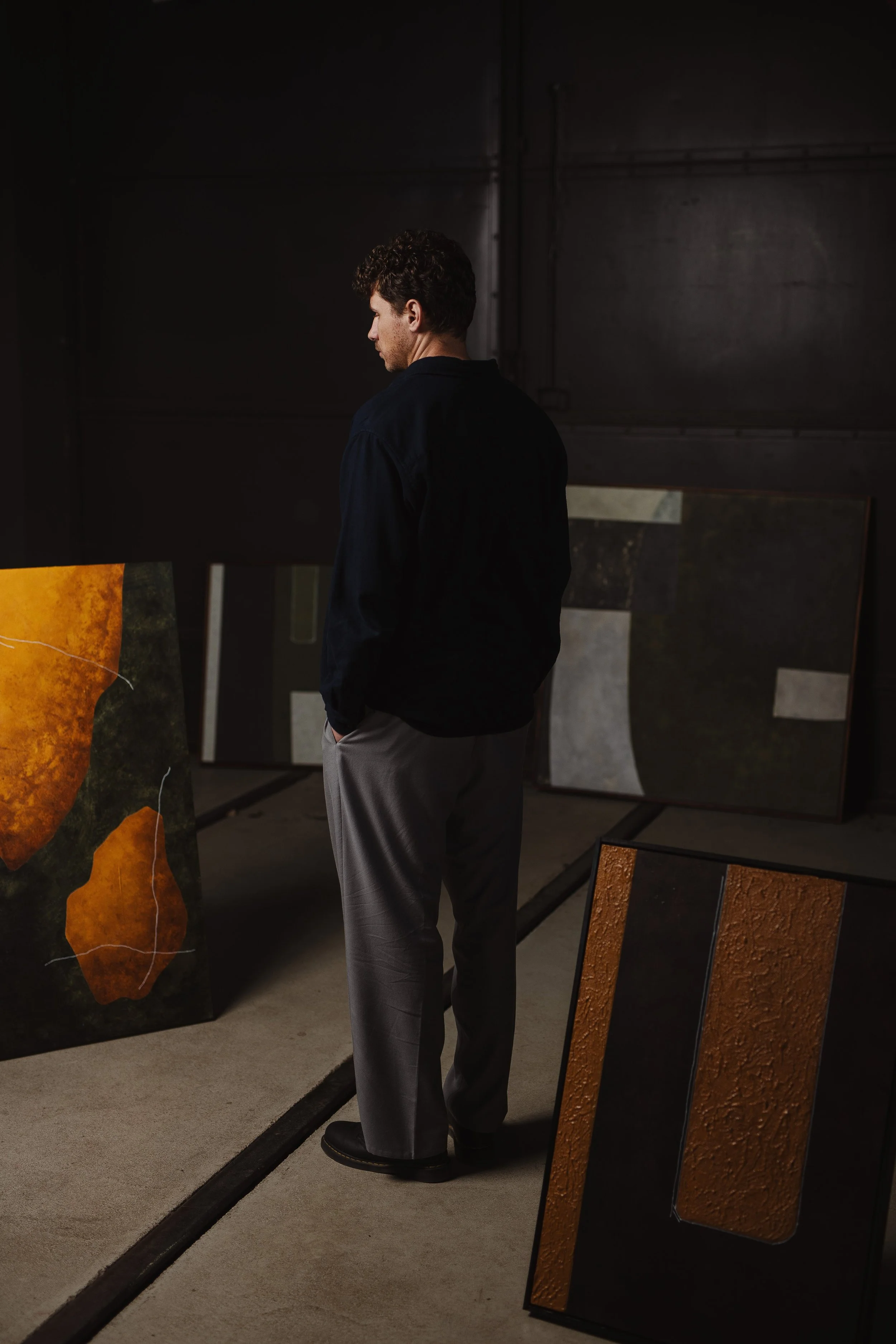 Ein junger Mann mit dunklen, lockigen Haaren trägt eine dunkle Jacke und graue Hosen. Er steht in einem dunklen Raum mit Kunstwerken und Gemälden aus verschiedenen Materialien, darunter große Leinwände mit abstrakten Mustern in warmen und erdigen Far