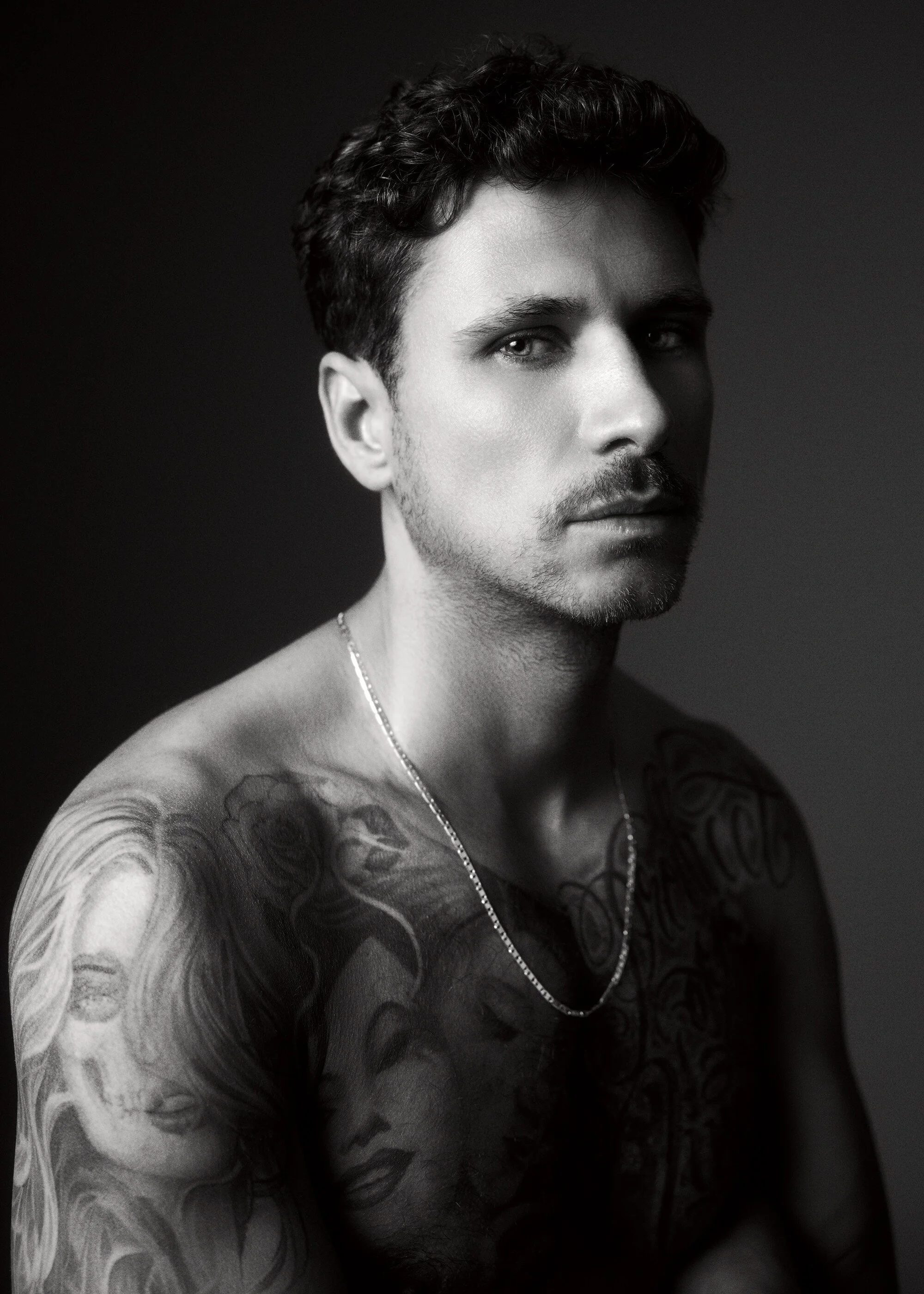 Schwarz-weiß-Fotografie eines jungen Mannes mit Tattoos auf Arme und Brust, kurzer dunkler Haartyp, Blick zur Kamera, ohne Hemd, trägt eine Halskette, dunkler Hintergrund.