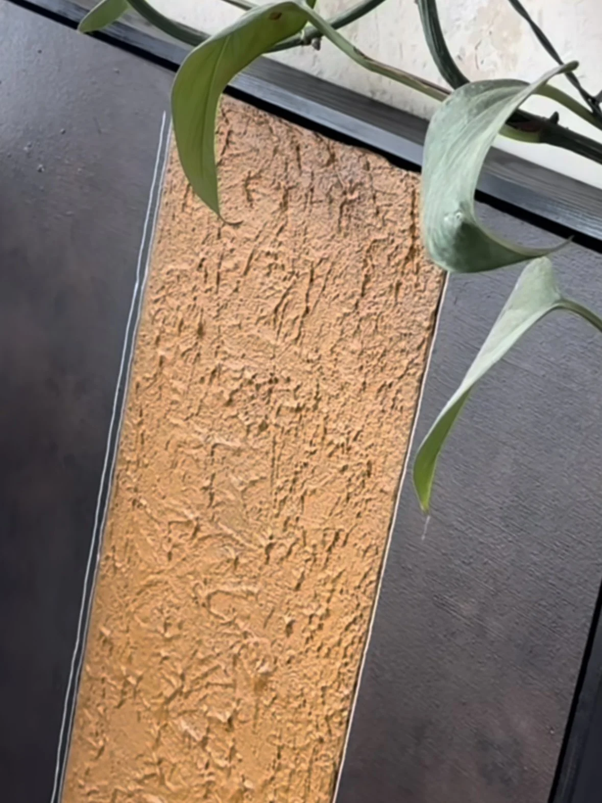 Blick auf eine vertikale orange strukturierte Wand mit grünen Pflanzen, die von oben und seitlich herabhängen.