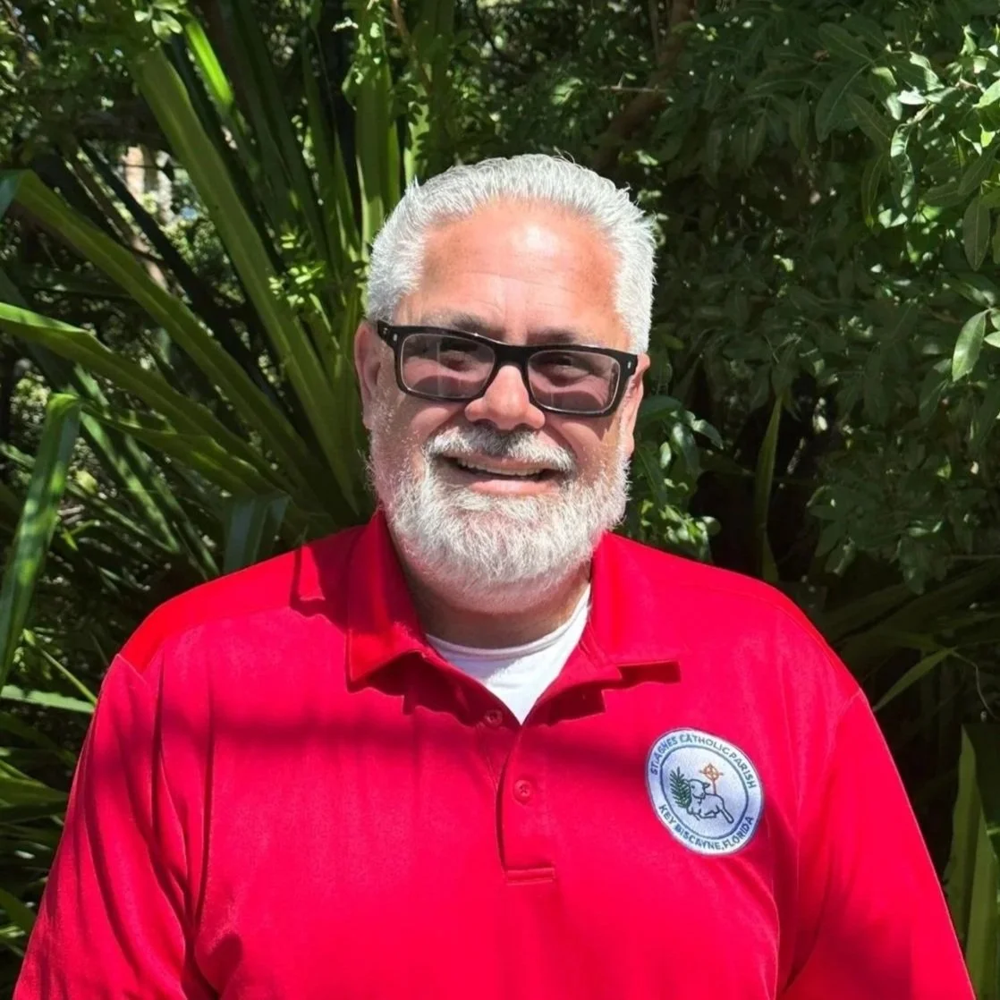 An older man with white hair, beard, and glasses, wearing a red polo shirt with a church emblem, standing outdoors with green plants in the background.