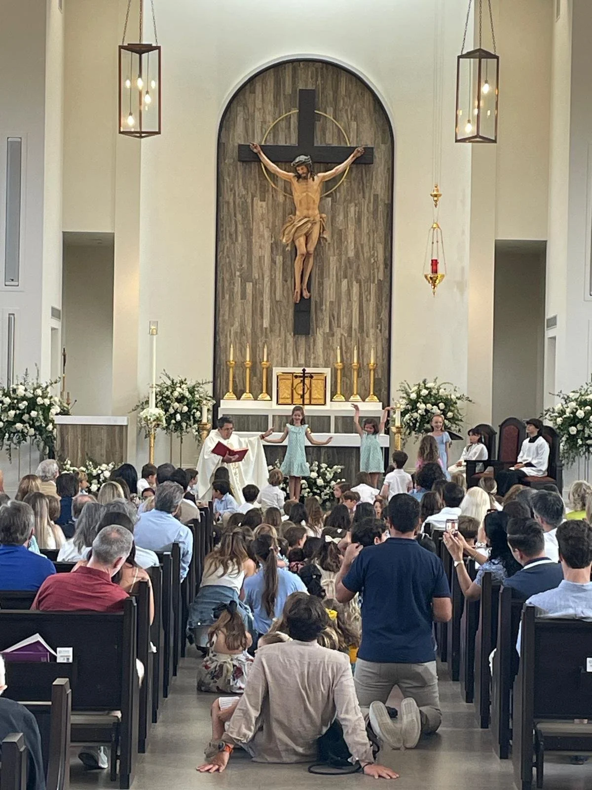 Children and a priest participating in a religious ceremony inside a church, with the altar and a crucifix with a depiction of Jesus Christ hanging above.