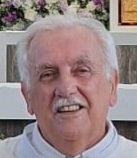 A close-up photo of an elderly man with white hair, smiling, against a background with a framed picture and furniture.