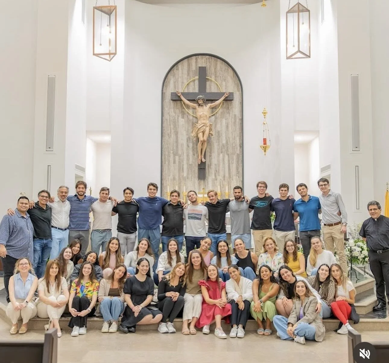 Group of young adults and older men posing in a church or chapel with an altar and a crucifix in the background.