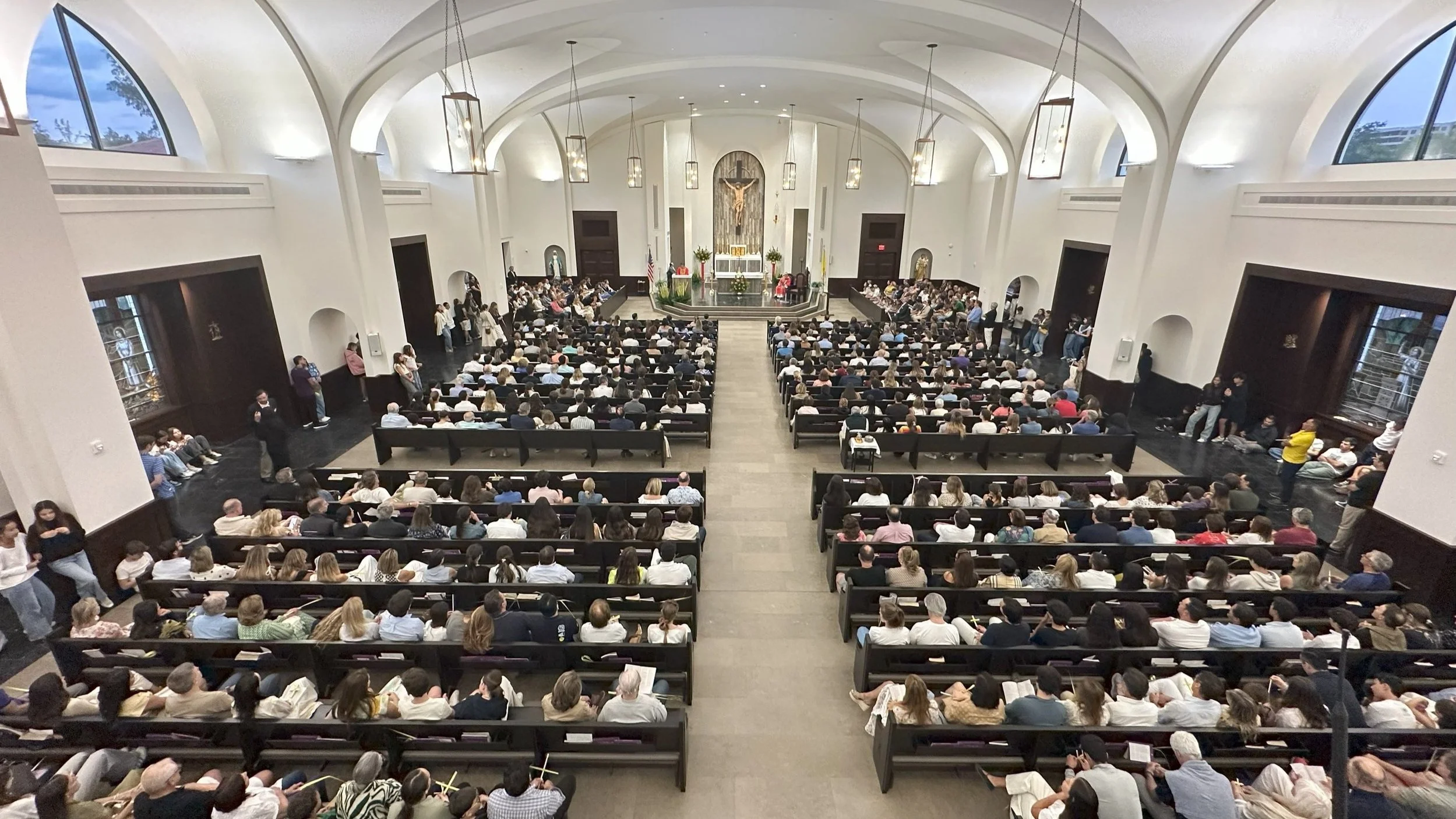 Inside a church with a congregation seated in pews, facing the altar with crucifix, decorated with flowers, during a service or event.