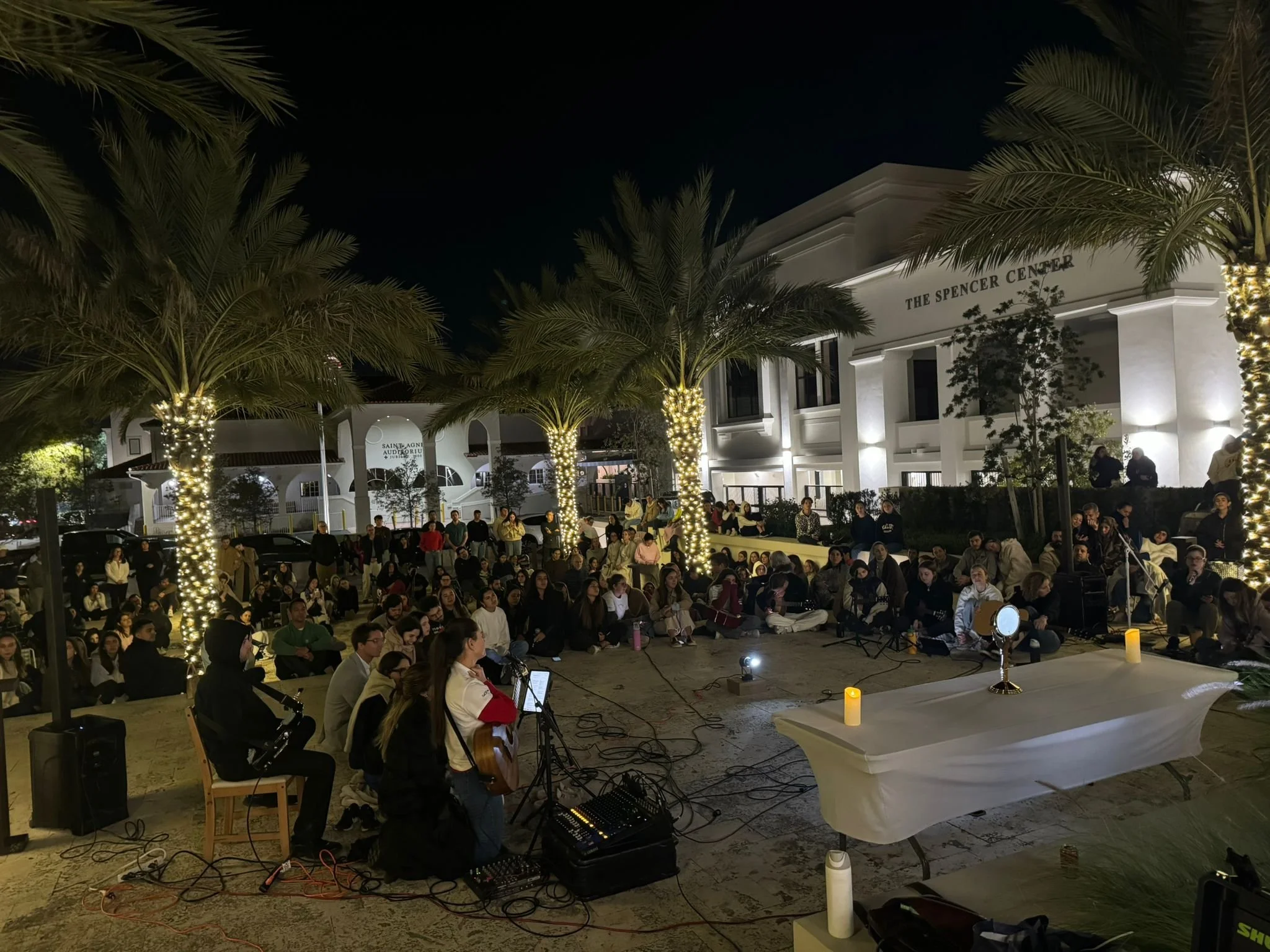 Outdoor night gathering with people sitting on steps, palm trees wrapped in string lights, and a stage with acoustic guitars and microphones in front of a white building labeled 'The Spencer Center'.