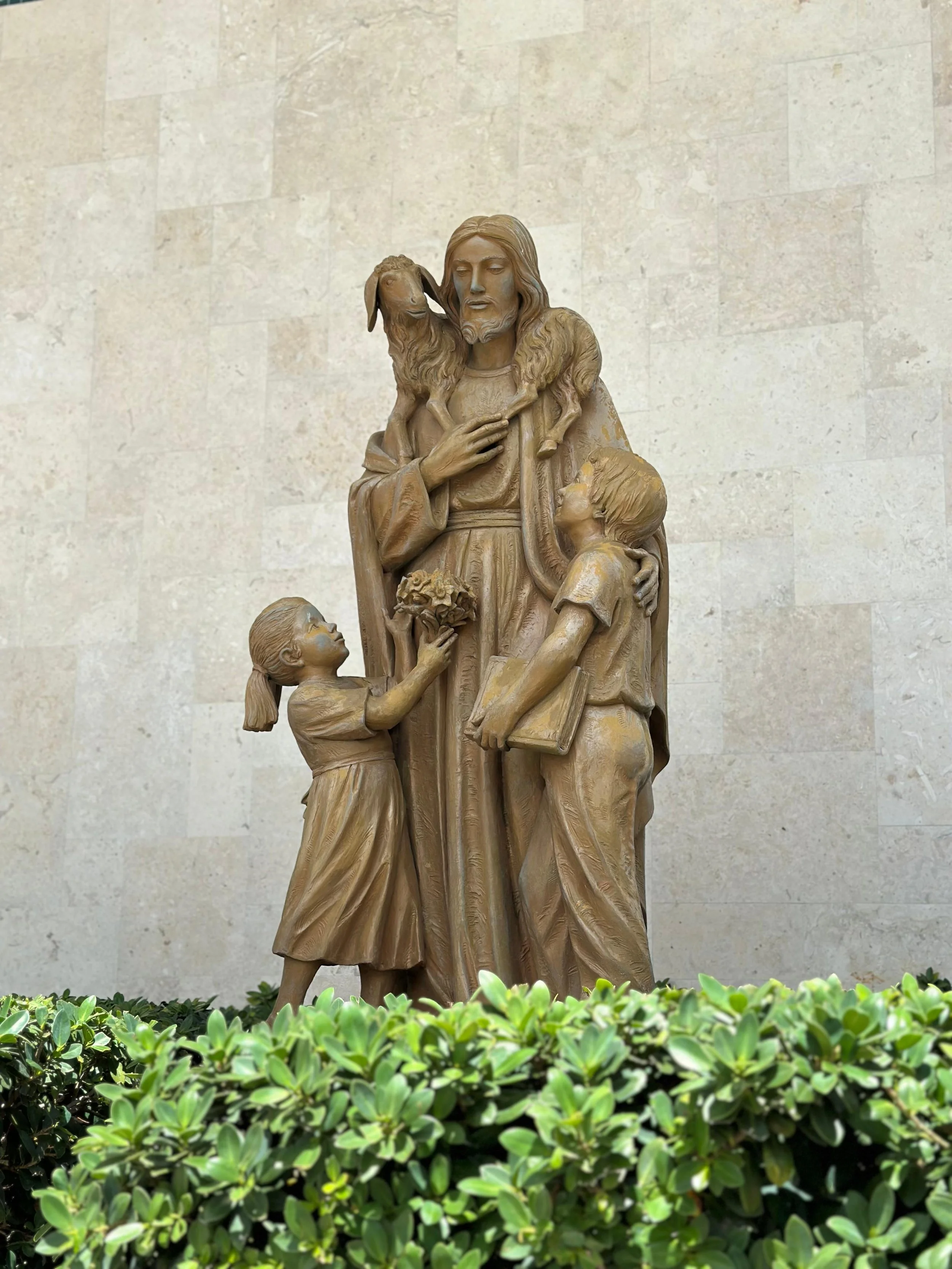 Wooden religious sculpture of Jesus Christ with children and a small lamb on his shoulders, standing in front of a beige stone wall and surrounded by green leafy plants.