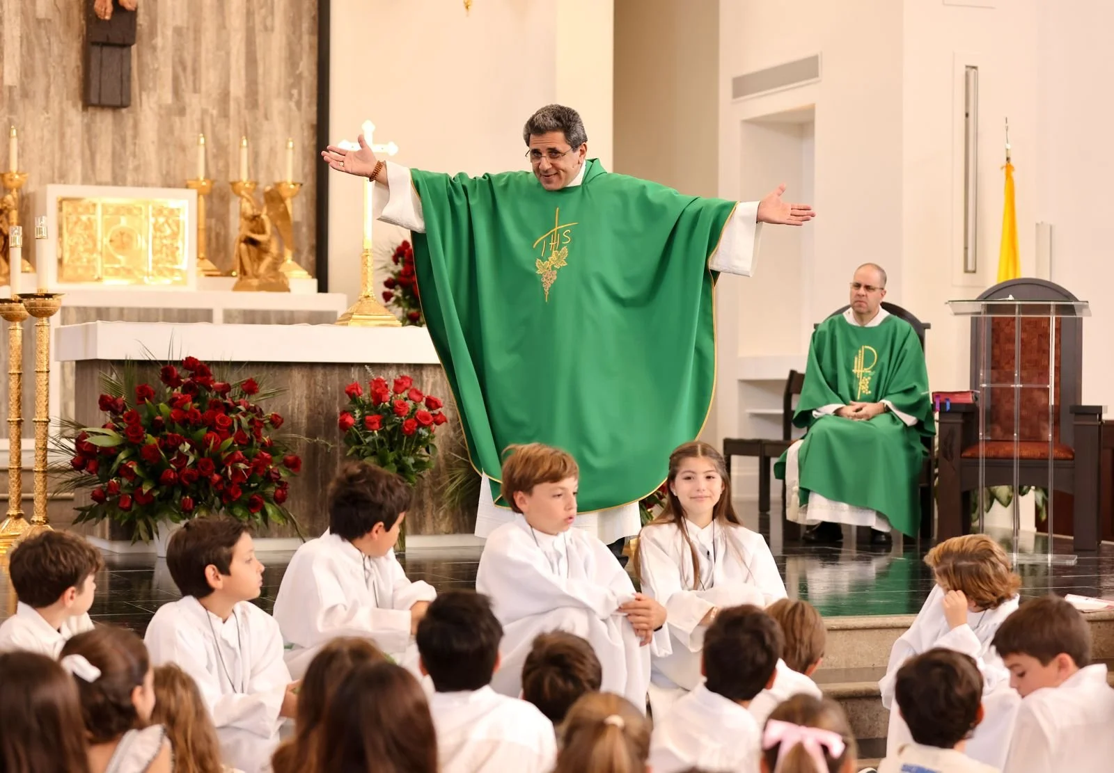 A priest with arms outstretched wearing a green vestment stands at the altar, surrounded by children in white robes. Another priest sits in the background. The altar is adorned with gold candles, a crucifix, and bouquets of red roses.