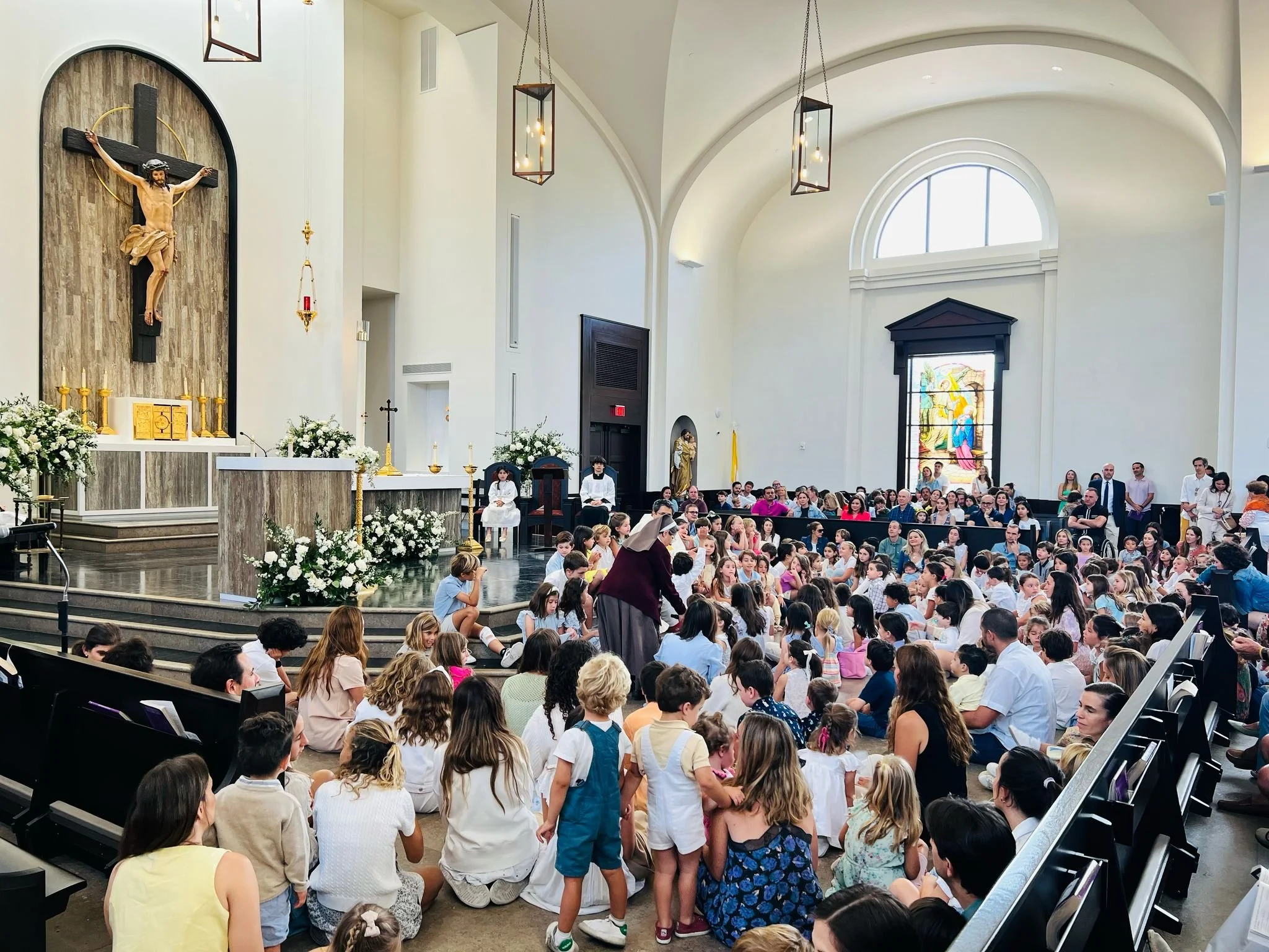 A church interior filled with children and adults seated on the floor and benches, with an altar, crucifix, and stained glass windows in the background.