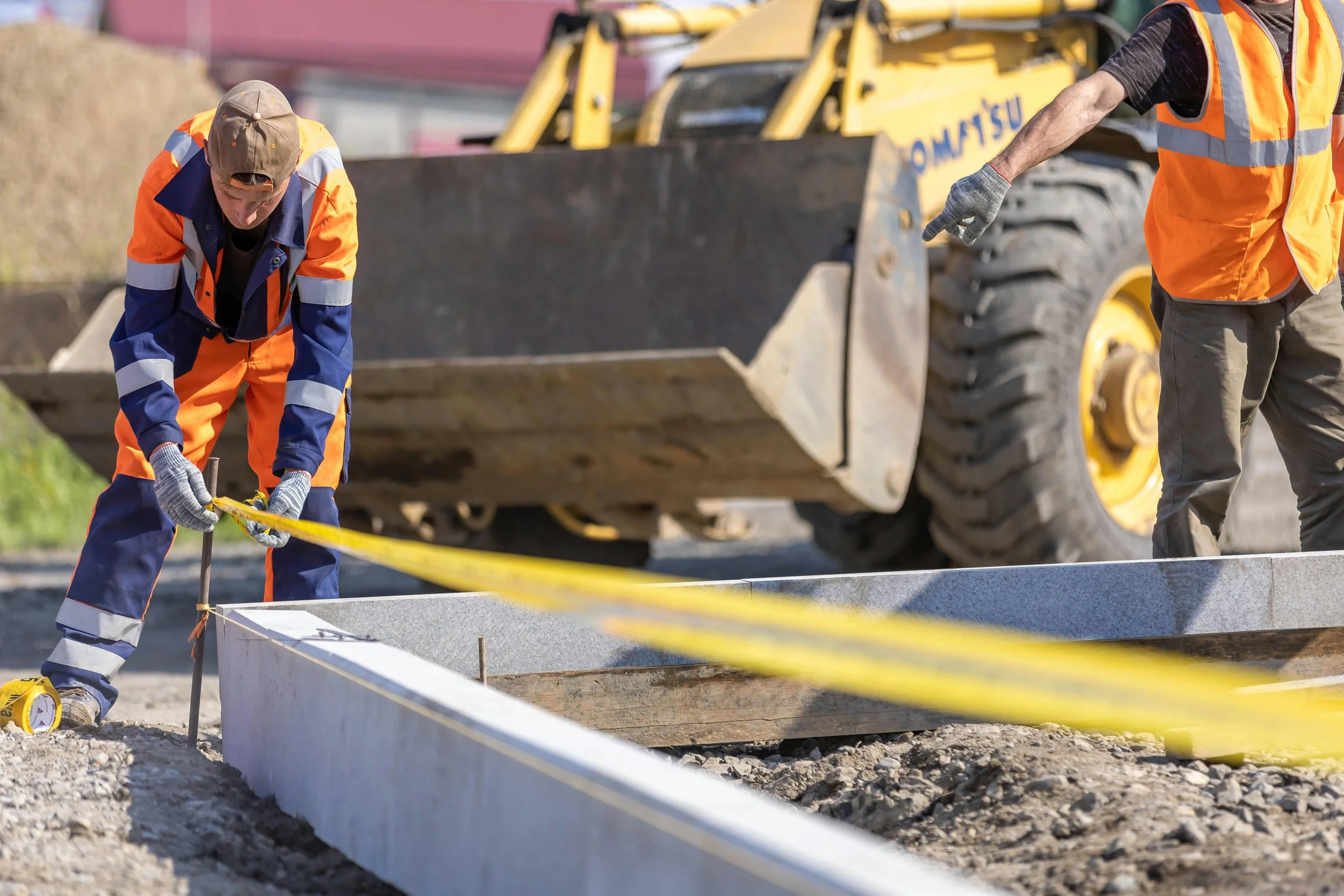 Trabajadores de construcción midiendo y colocando un marco de concreto en una obra en construcción con maquinaria pesada en el fondo.