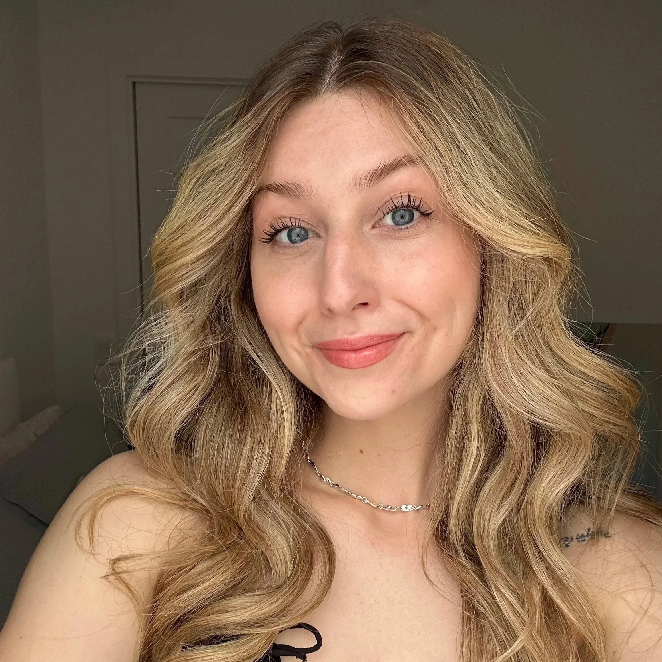 Close-up of a woman with wavy blonde hair, blue eyes, light makeup, and a silver necklace, smiling slightly, with a blurred indoor background.
