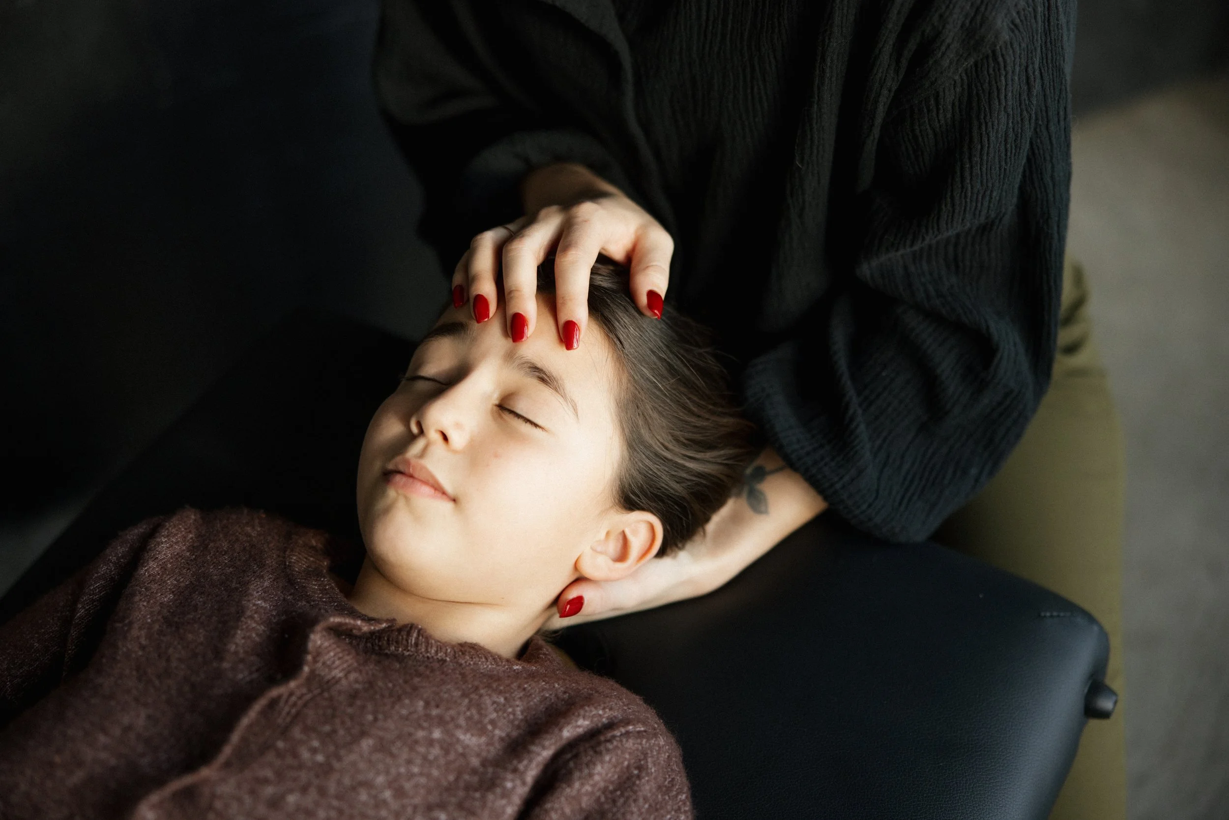A person with red nails gently holding a young girl's head while she lies peacefully with closed eyes, receiving a craniosacral fascial therapy treatment.