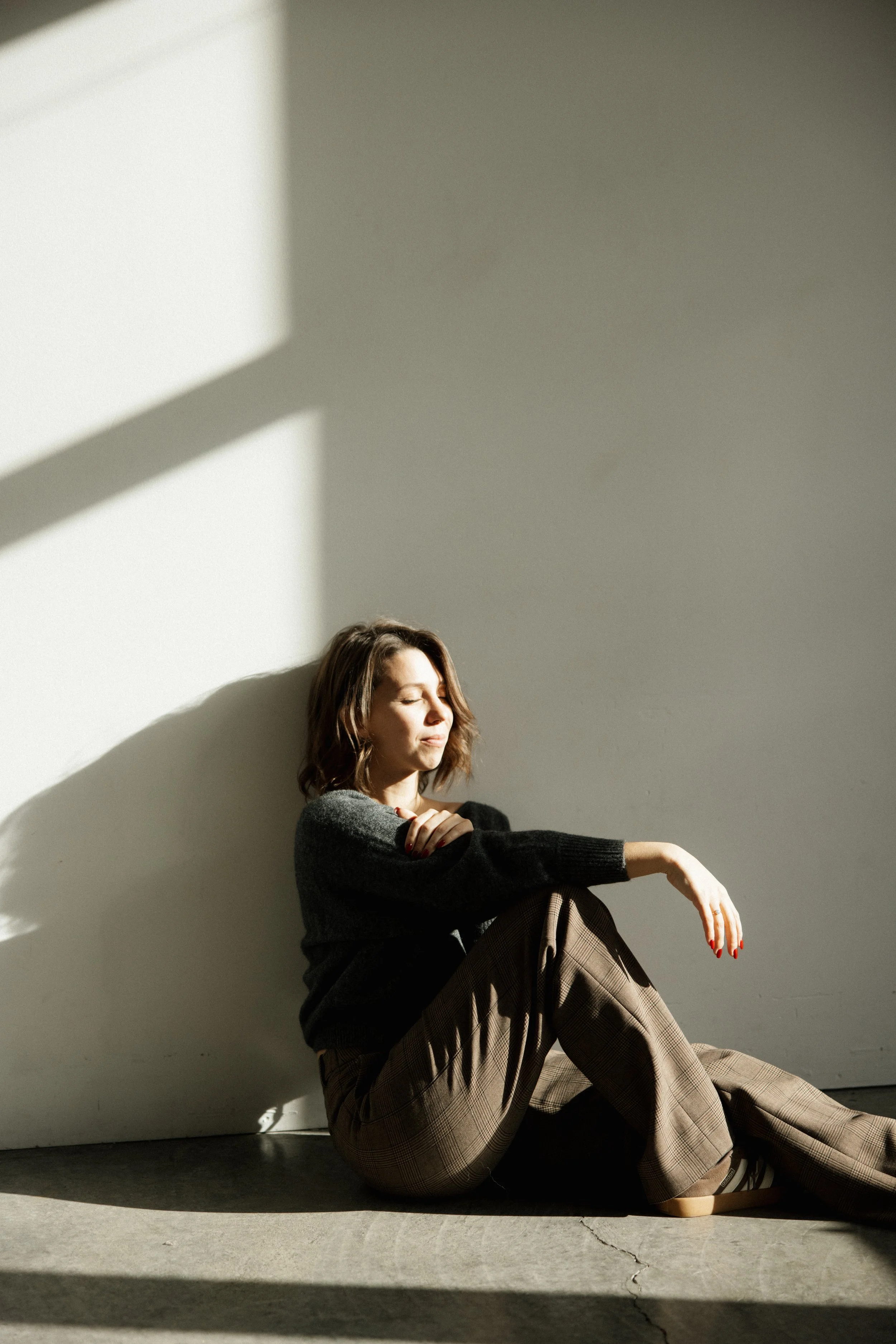 A woman sitting on the floor in a minimal room, with sunlight casting shadows on the wall behind her, eyes closed, wearing a dark sweater and plaid pants.