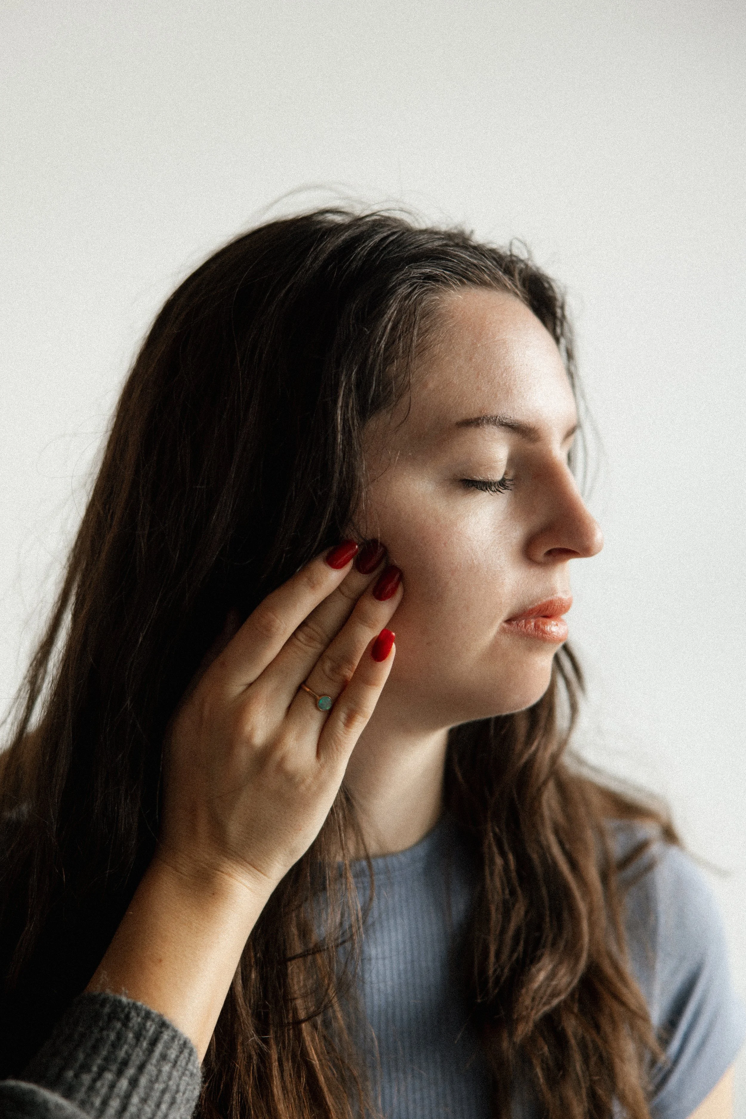 A woman with long brown hair has her eyes closed and touches her cheek with her hand, which has red nails and a ring with a turquoise stone who is doing CFT