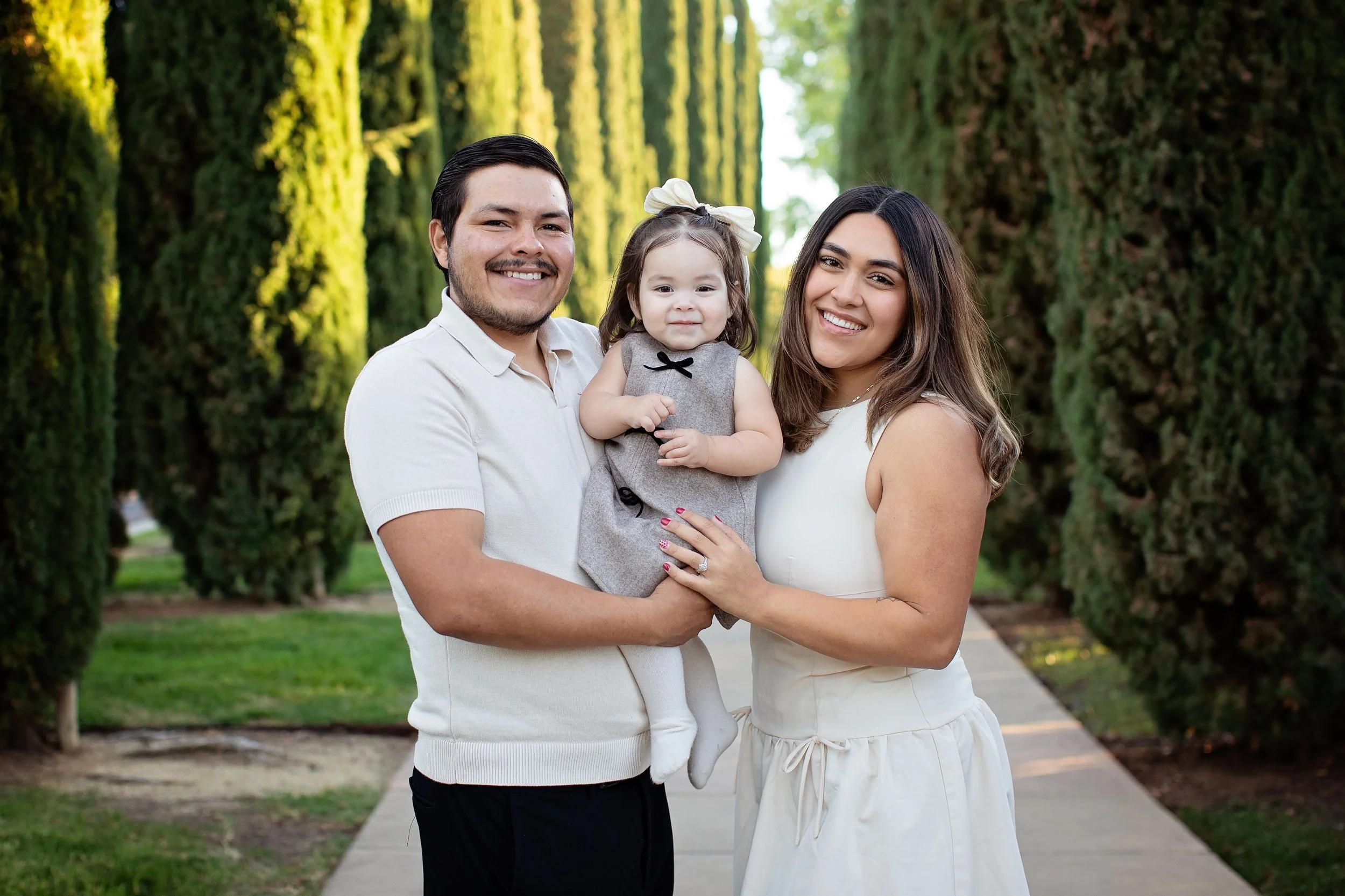 A family of three smiling and standing outdoors on a pathway surrounded by tall green trees.
