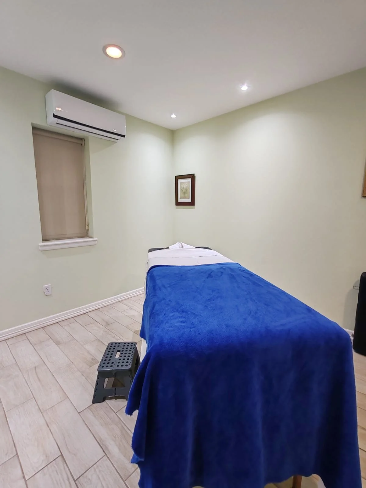 Sala de terapia con cama y silla pequeña, paredes pintadas de color verde claro, una ventana con cortina beige, un aparato de aire acondicionado en la pared, cuadros decorativos en las paredes, piso de baldosas de color beige.