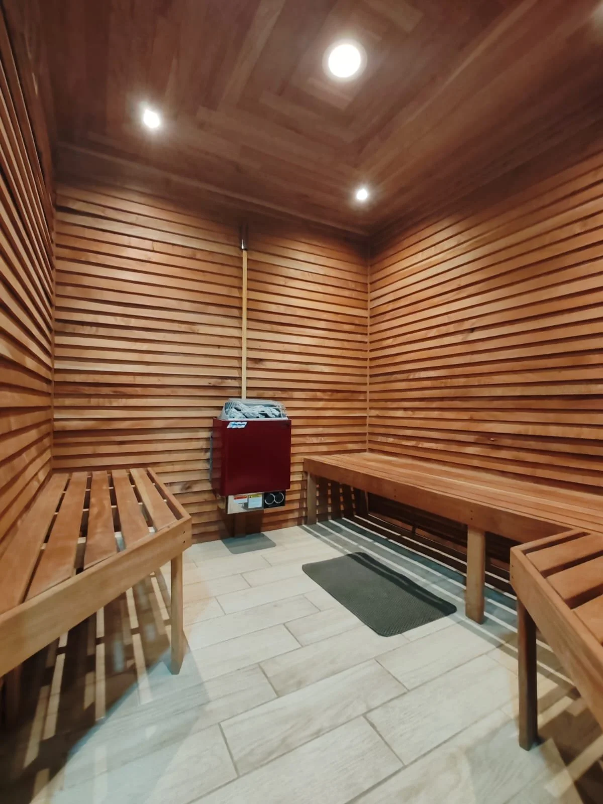 Sauna con paredes y techo de madera, bancos de madera en las esquinas y una alfombra pequeña en el suelo. En la pared, un calefactor portátil y una lámpara en el techo.