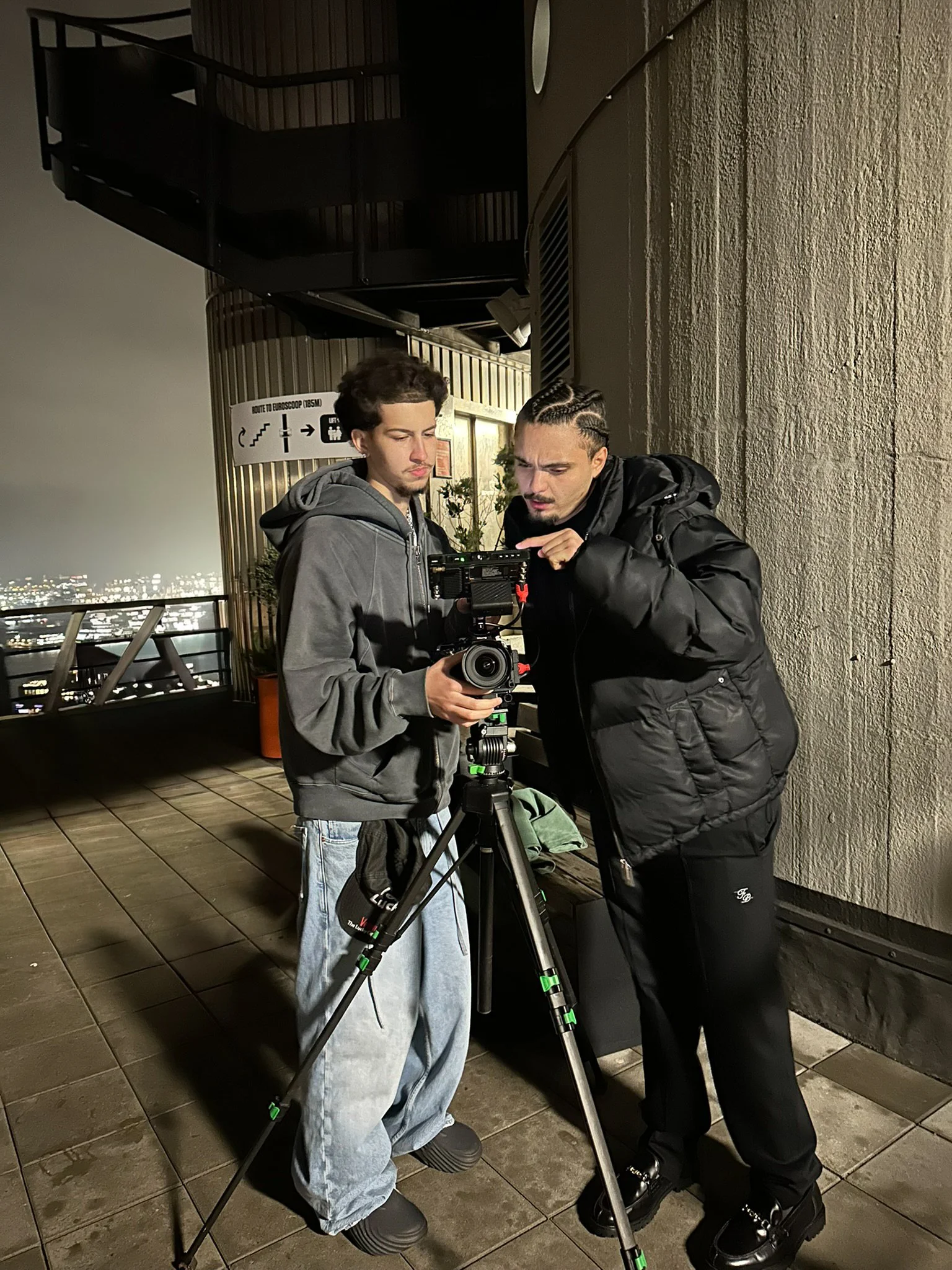 Two men working with a camera on a tripod on a rooftop at night, with city lights visible in the background.