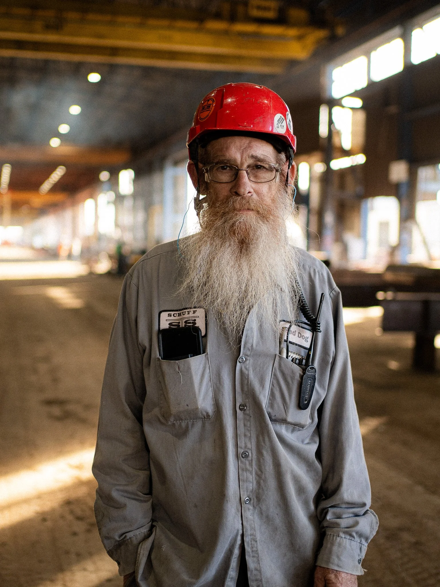 A man with glasses and a long gray beard wearing a red safety helmet and gray work coveralls standing inside a construction or industrial building with a dirt floor and large windows in the background.