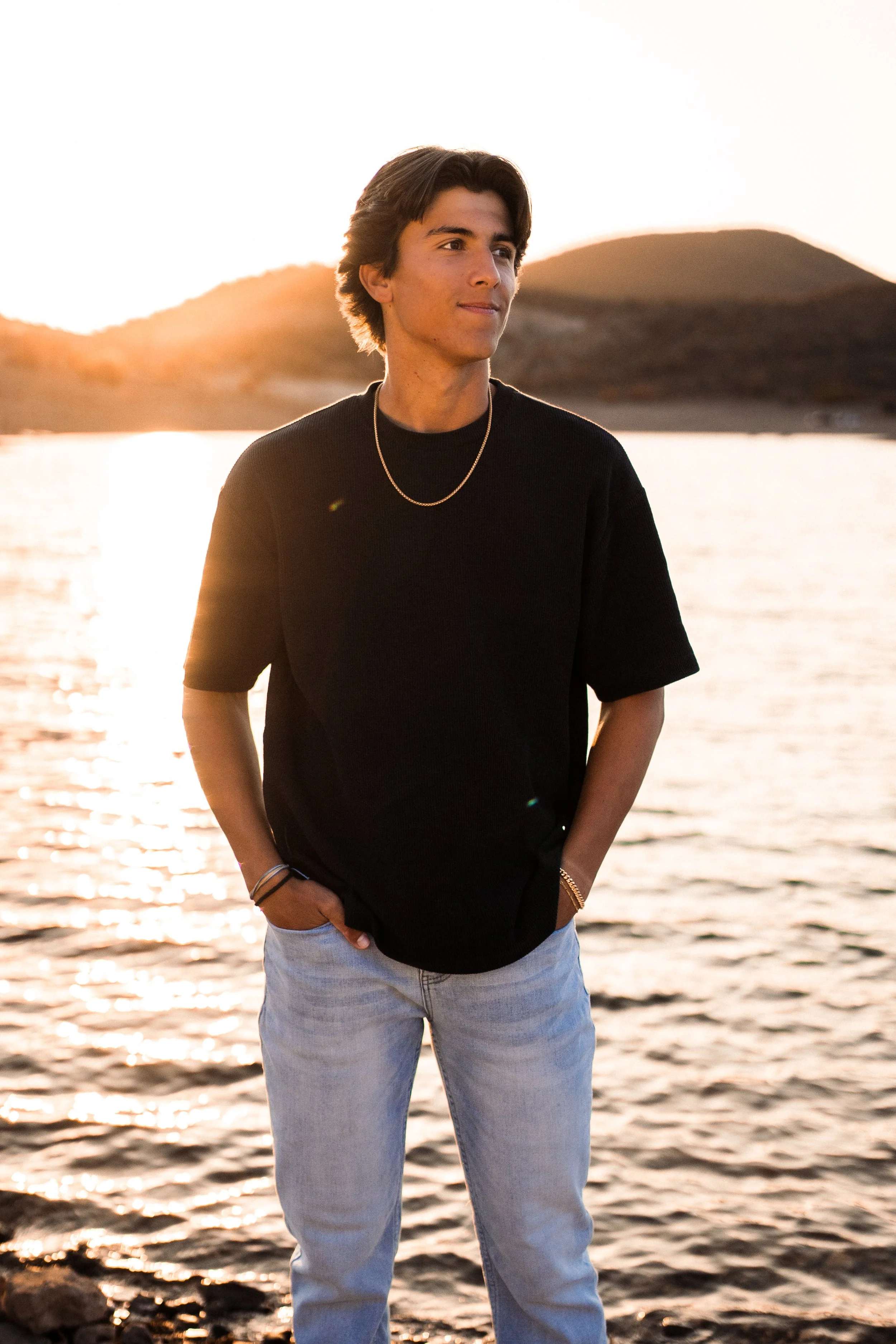 A young man stands by a lake at sunset, wearing a black shirt and light jeans, with his hands in his pockets, looking to the side.