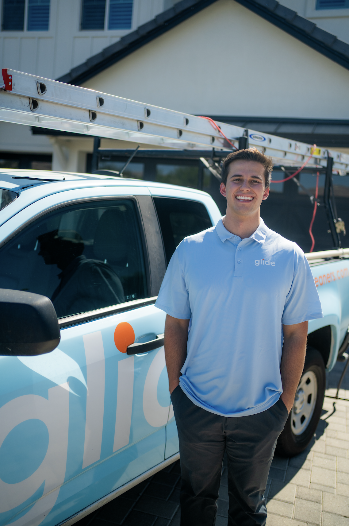 A smiling young man in a light blue shirt standing next to a company branded service truck with a ladder on top.